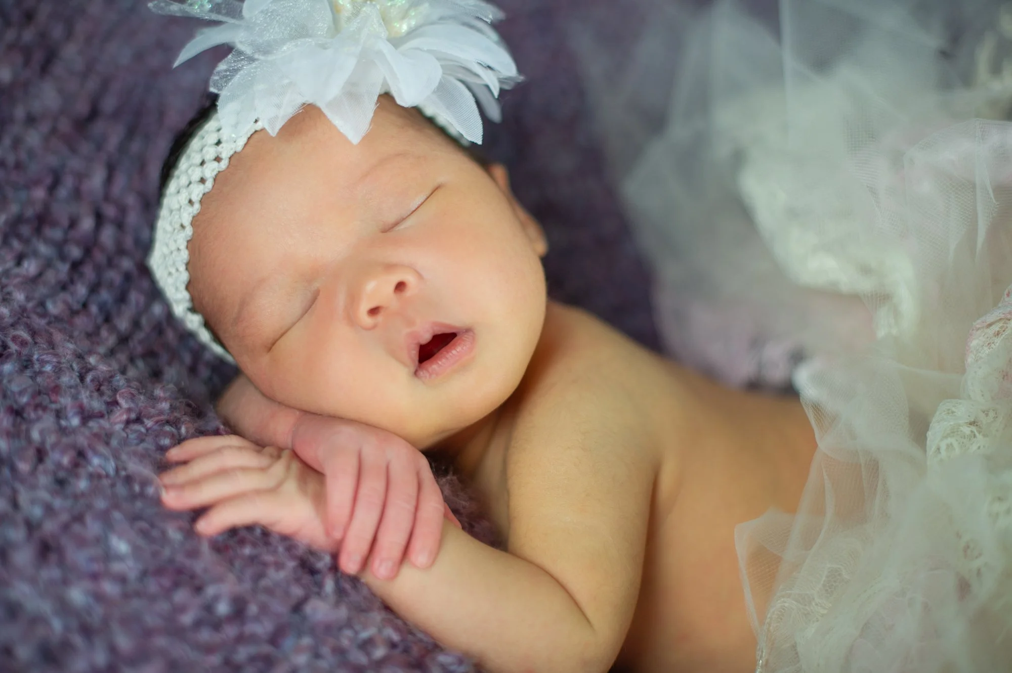 Newborn baby sleeping, wearing a white headband with a large flower, wrapped in a soft, purple blanket.