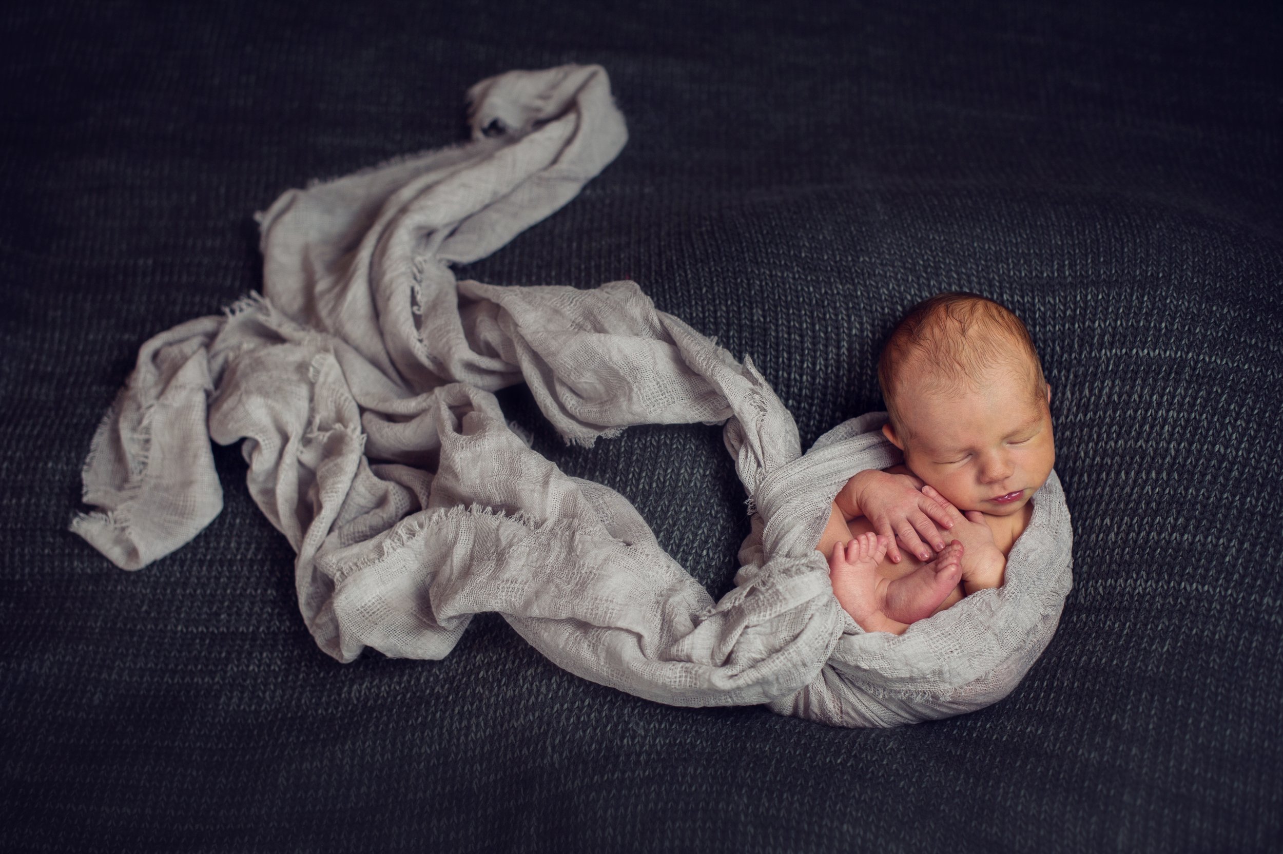 Newborn baby wrapped in a soft, gray blanket sleeping peacefully on a dark textured surface.