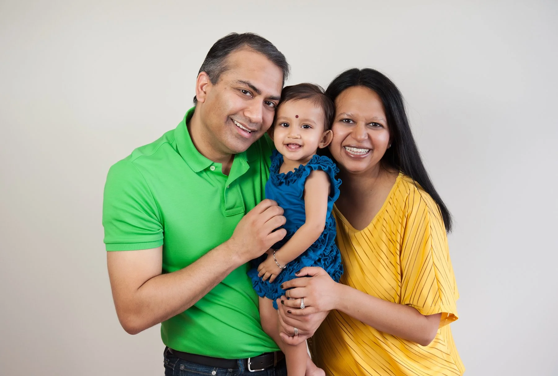 A studio portrait of a smiling family of three with a man in a green shirt, a woman in a yellow shirt, and a young child in a blue dress, standing against a plain background.