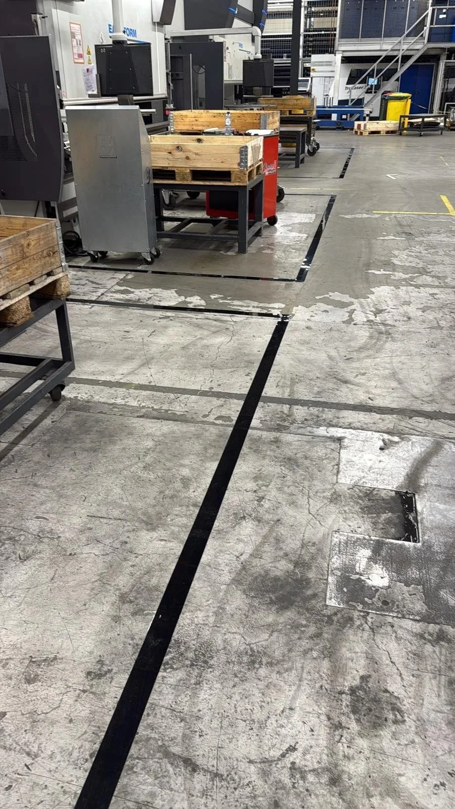 Industrial workspace with machinery, wooden crates, and black floor markings on a concrete floor in a factory or manufacturing plant.