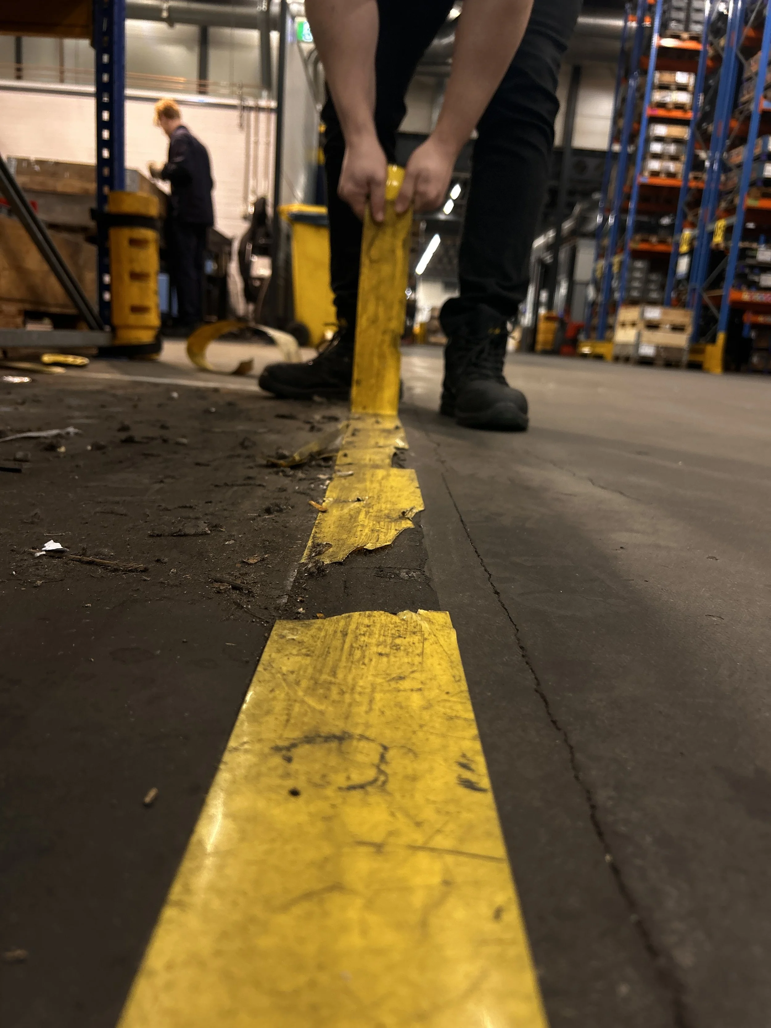 Worker in black boots and black pants applies yellow floor marking tape in a warehouse.