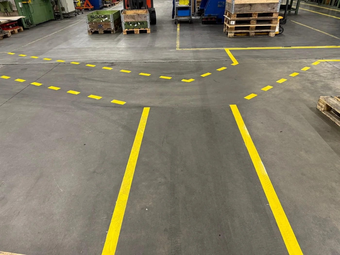 Yellow lines and markings on concrete floor indicating designated areas in an industrial or warehouse setting, with pallets and equipment in the background.