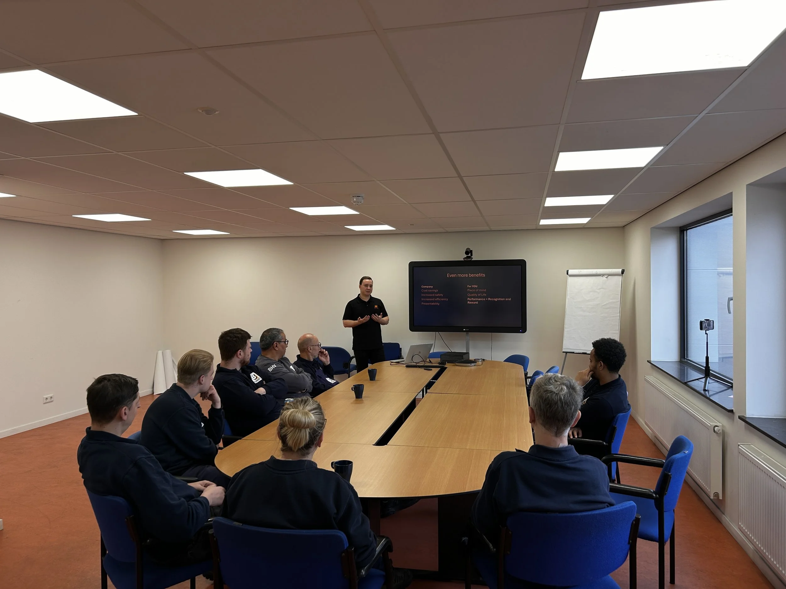 Magnus is giving a presentation in a conference room with several operators seated around a large U-shaped table, facing a screen displaying a PowerPoint slide.