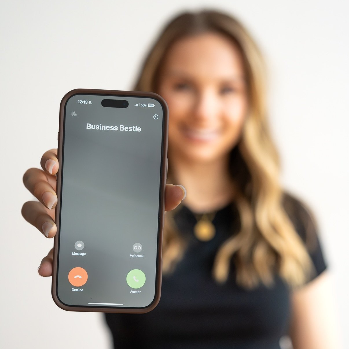 A woman with long blonde hair holding a smartphone displaying an incoming call from 'Business Bestie' and smiling.