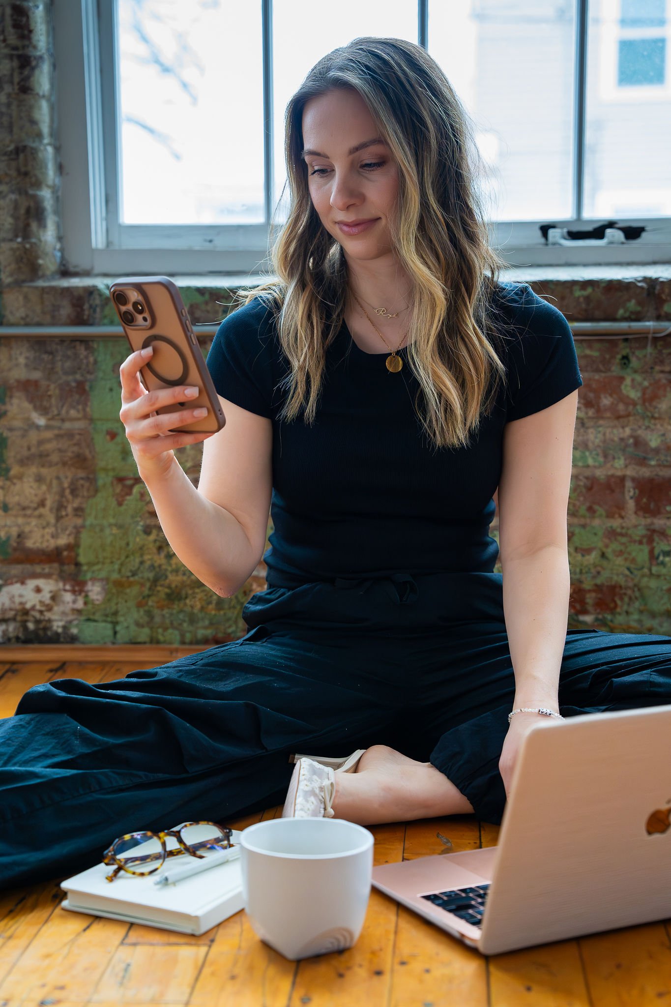 A woman with wavy hair sitting cross-legged on a wooden floor in front of a window, looking at her phone. She is wearing a black t-shirt and black pants, with a laptop, a notebook, a pair of glasses, and a white mug in front of her.