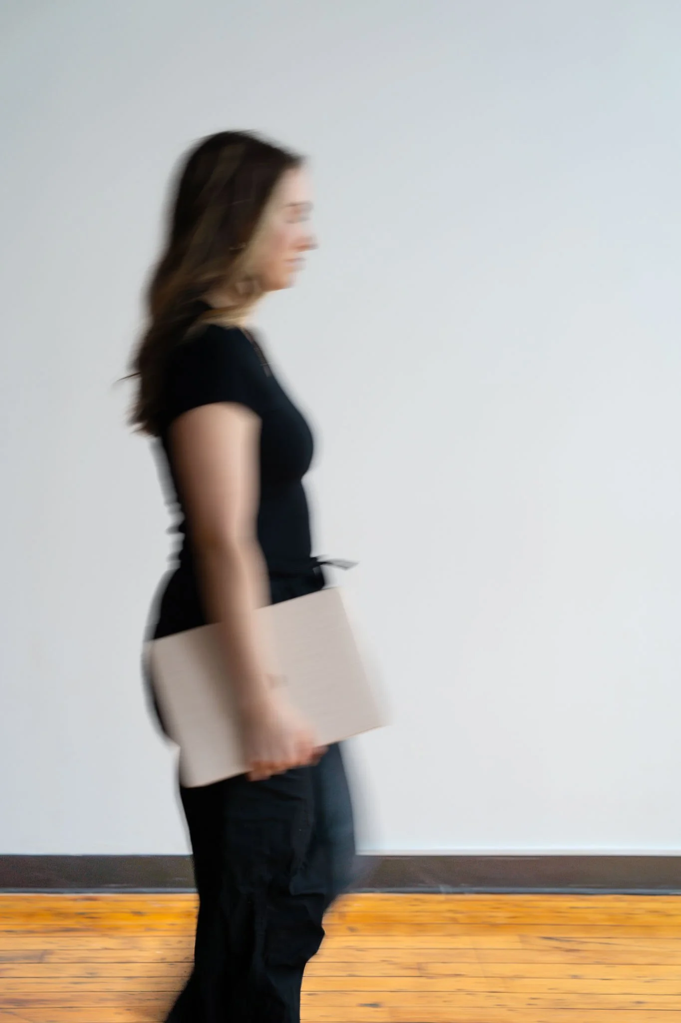 Blurred image of a woman standing sideways, holding a closed laptop, against a plain white wall and wooden floor.