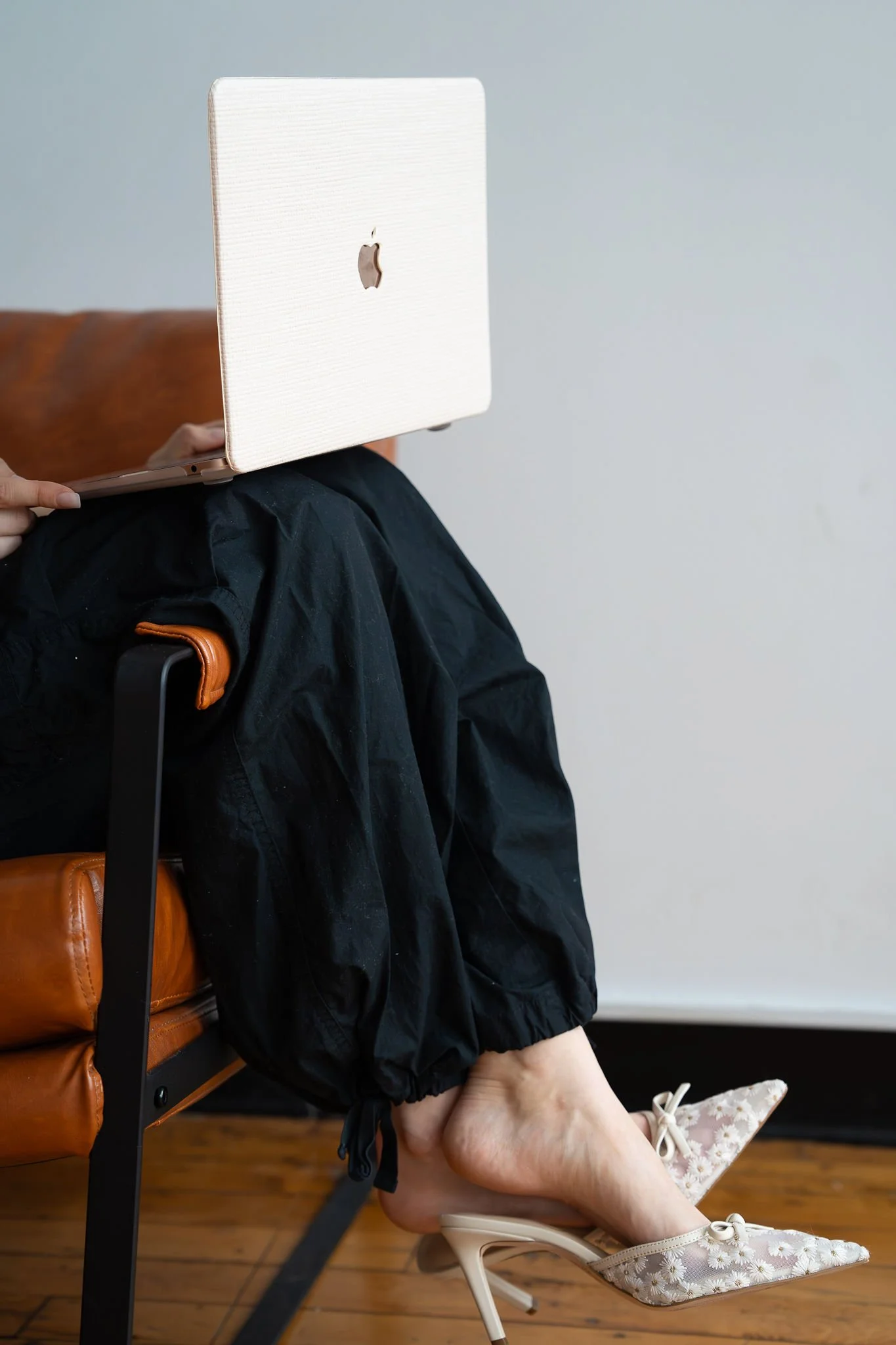 Person sitting on a chair with a white Apple MacBook on their lap, wearing black pants and beige floral high heels with pointed toes.