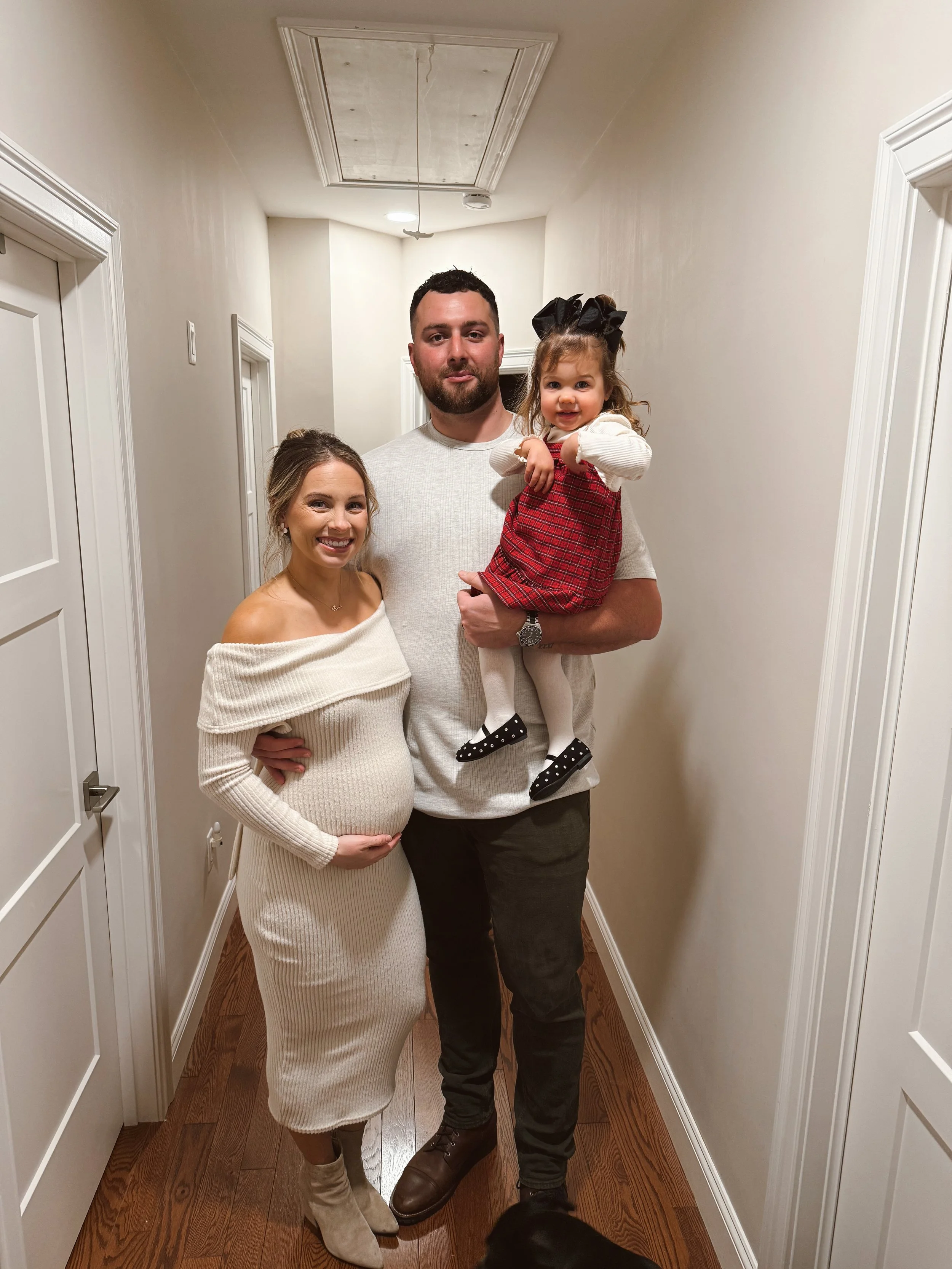 A family of three standing in a corridor. The woman is pregnant and wearing a cream off-the-shoulder knit dress, smiling and holding her belly. The man is wearing a light grey shirt and dark pants, holding a young girl. The girl is dressed in a red plaid dress with a white top, white tights, and black shoes with white polka dots, smiling and pointing.