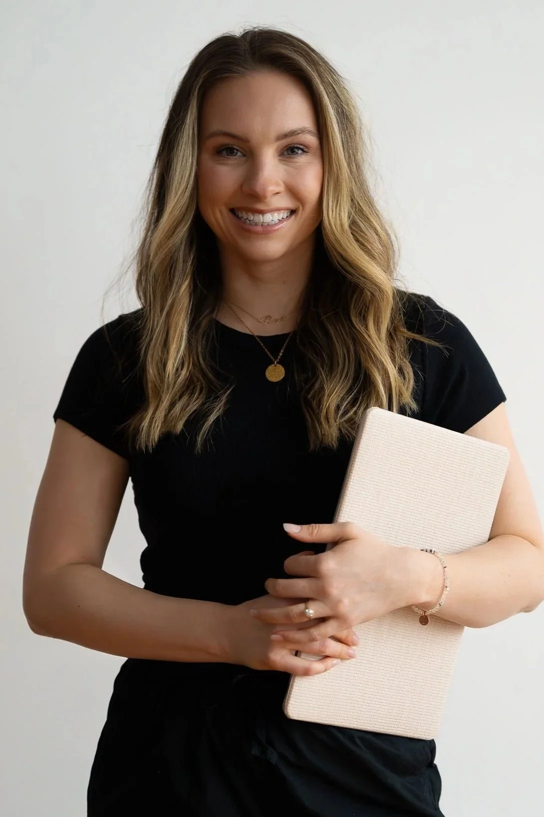 A young woman with wavy blonde hair is smiling and holding a light-colored notebook against a plain background.