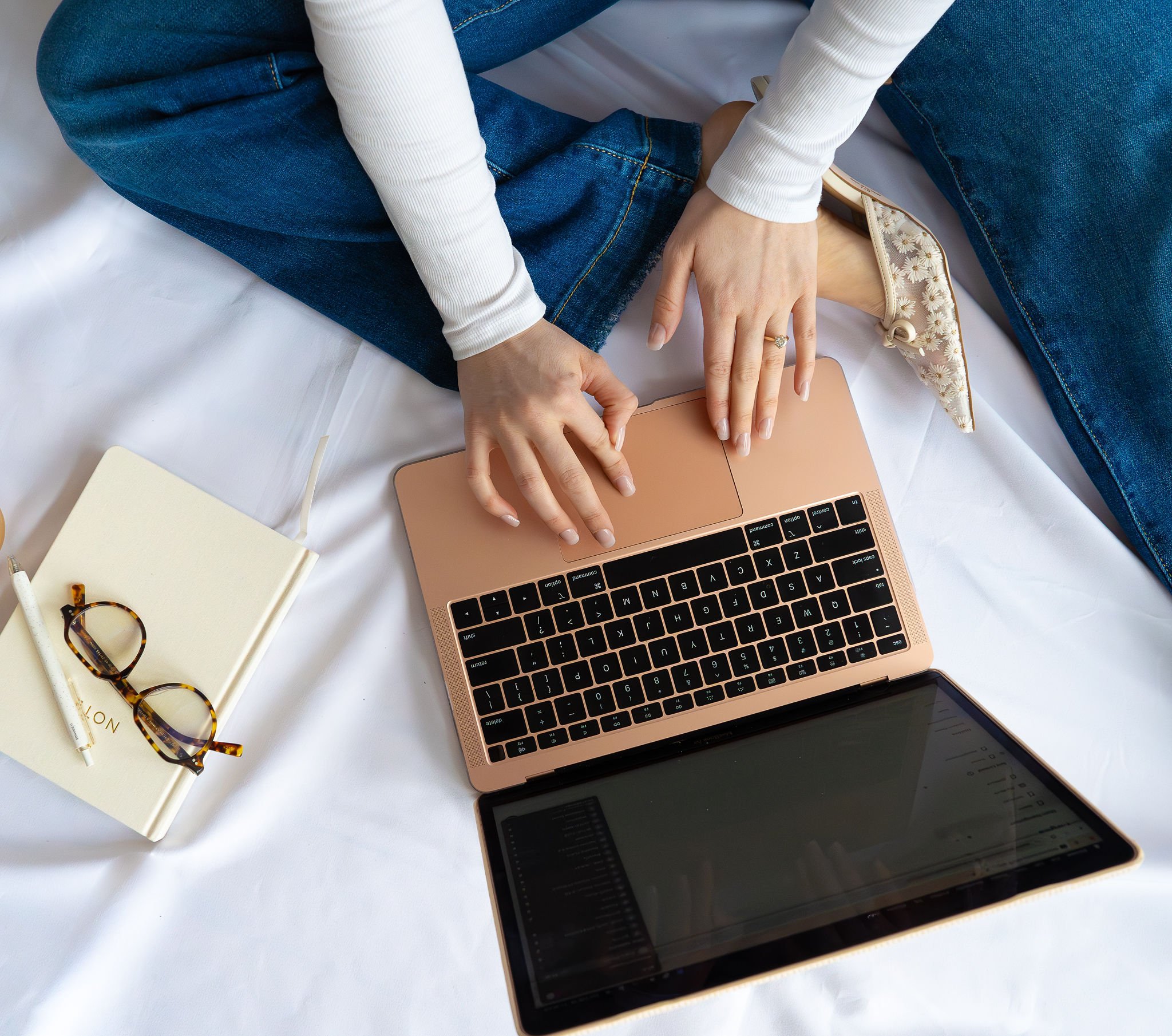 Person sitting on a white bed using a pink laptop, with glasses, a notebook, a pen, and a tablet nearby.