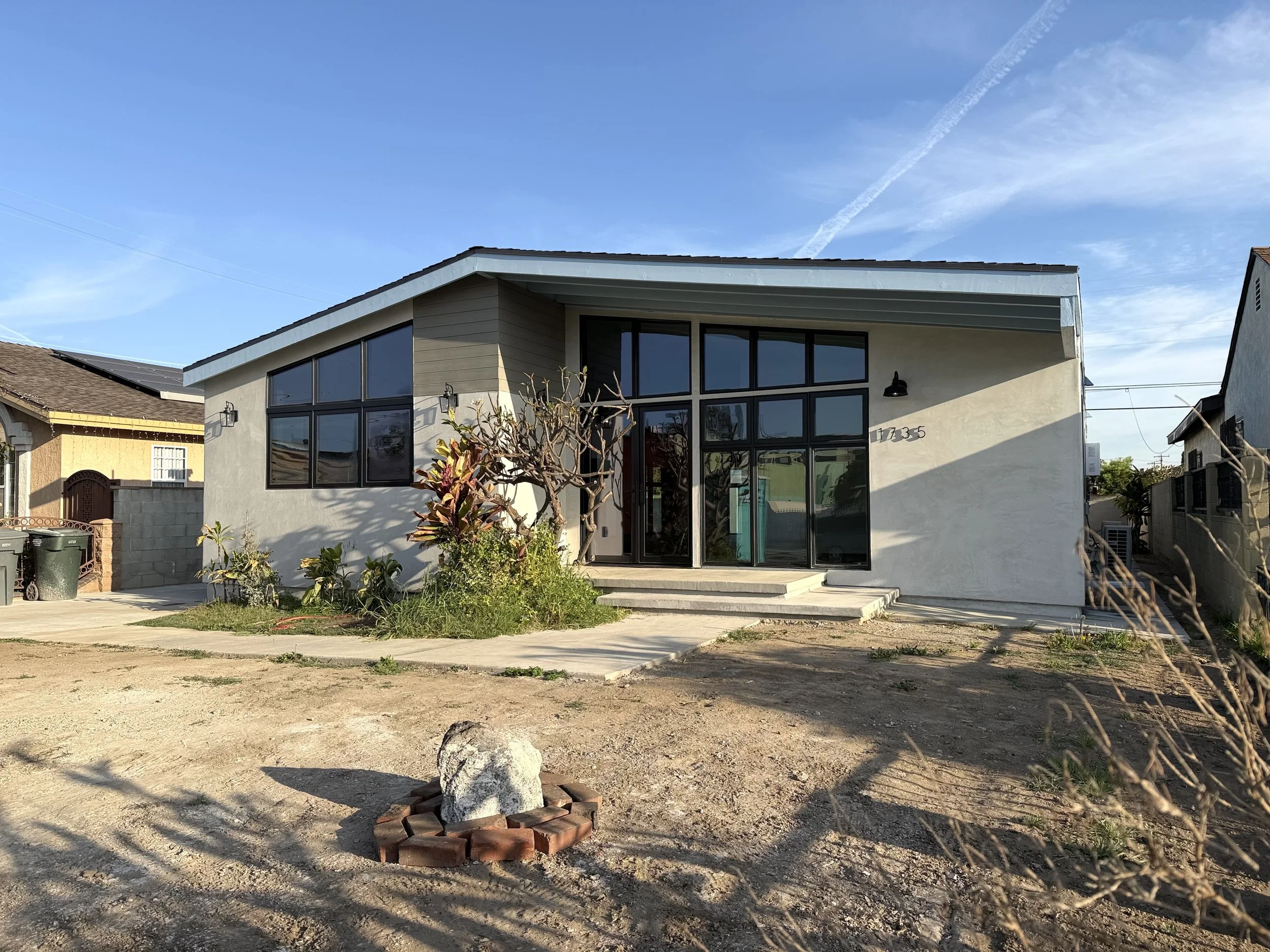 Modern house with large glass windows and front steps, surrounded by bare soil and a small garden with a leafless tree. Clear blue sky with clouds and airplane contrails in the background.