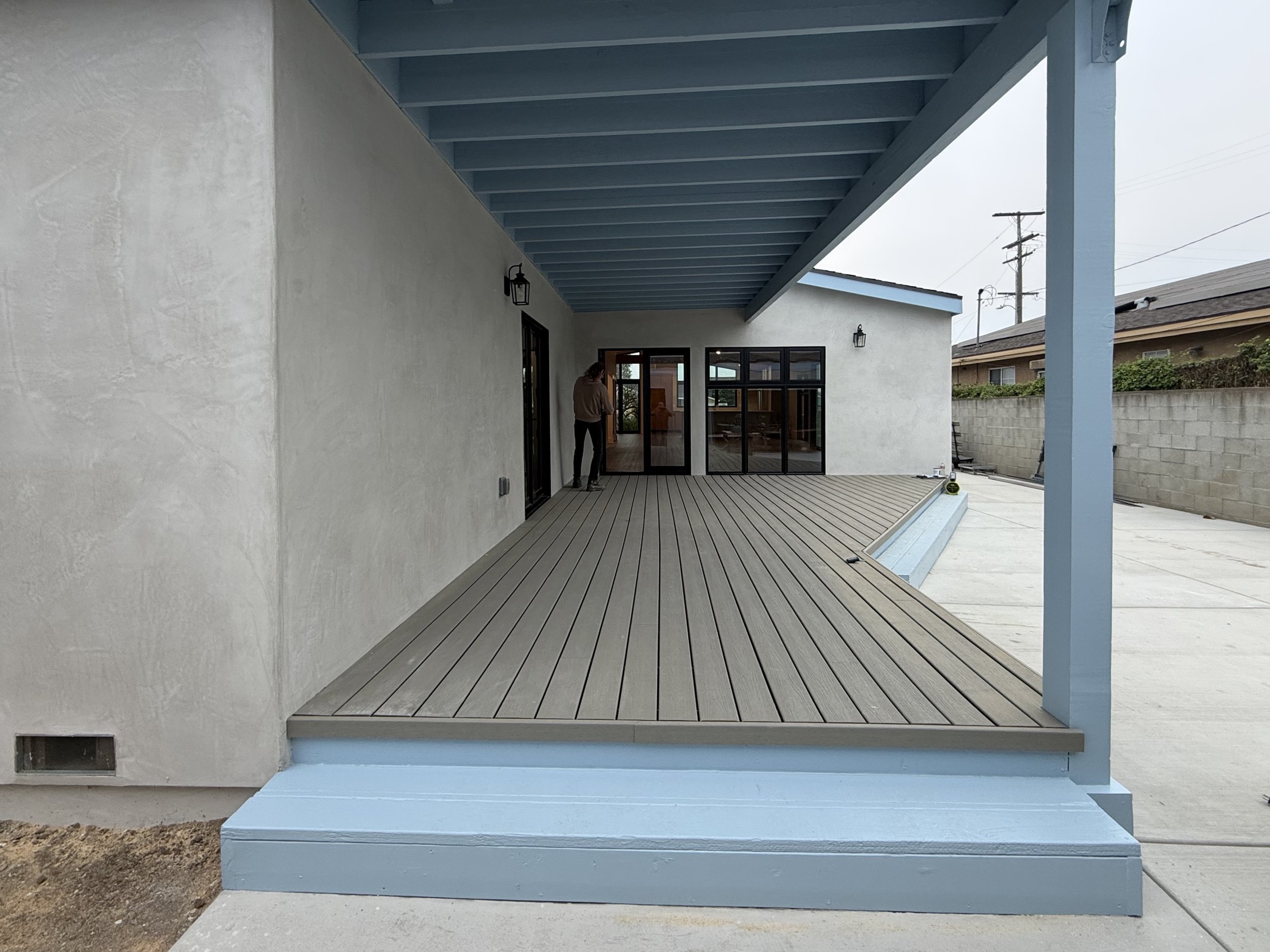 Modern house backyard with wooden deck porch, black-framed glass doors, and outdoor lighting fixtures, with a person standing near the doors.