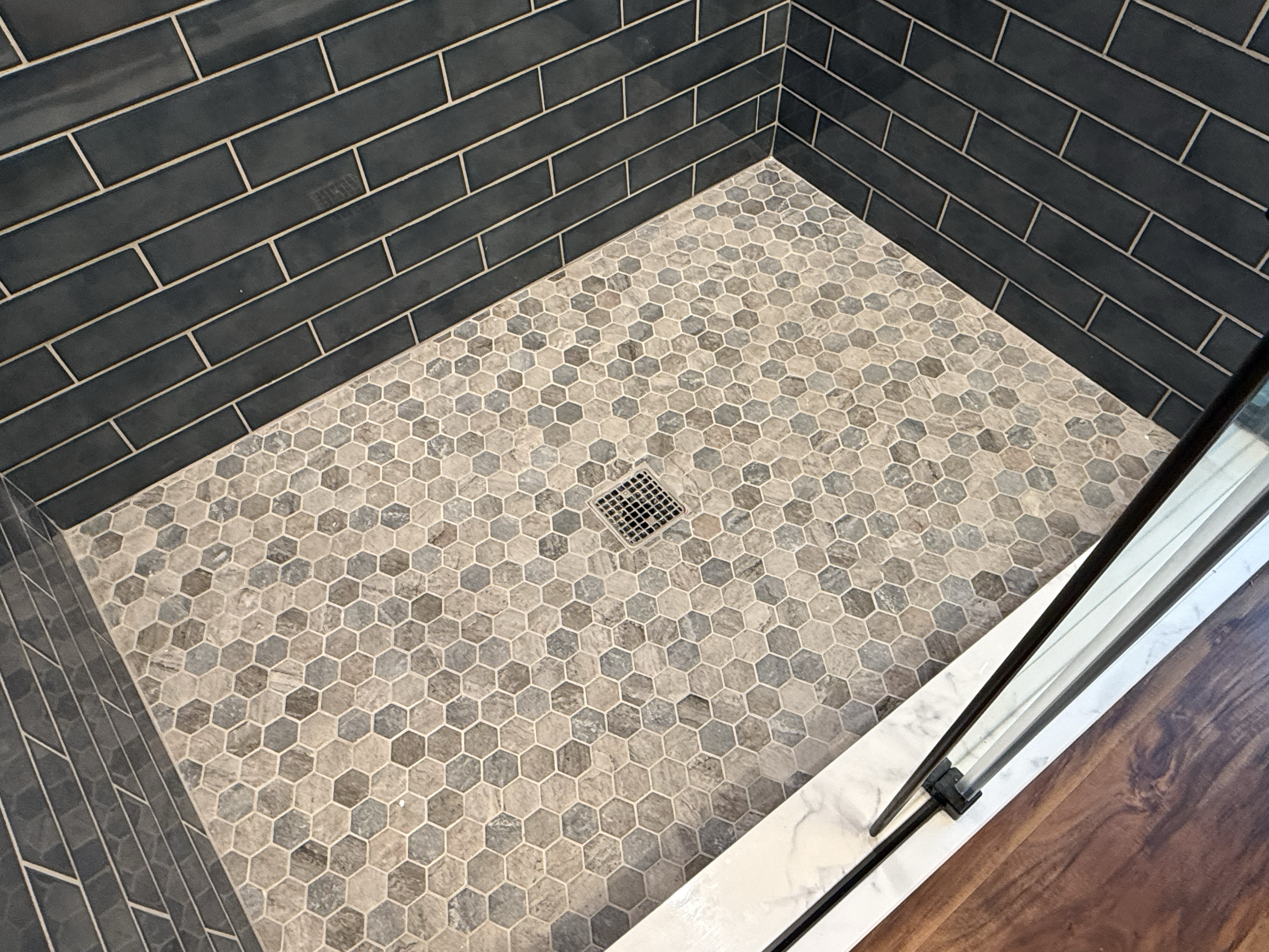 Empty shower area with hexagonal beige and gray tiles on the floor, black rectangular tiles on the walls, metal drain on the floor, and glass door with metal frame.