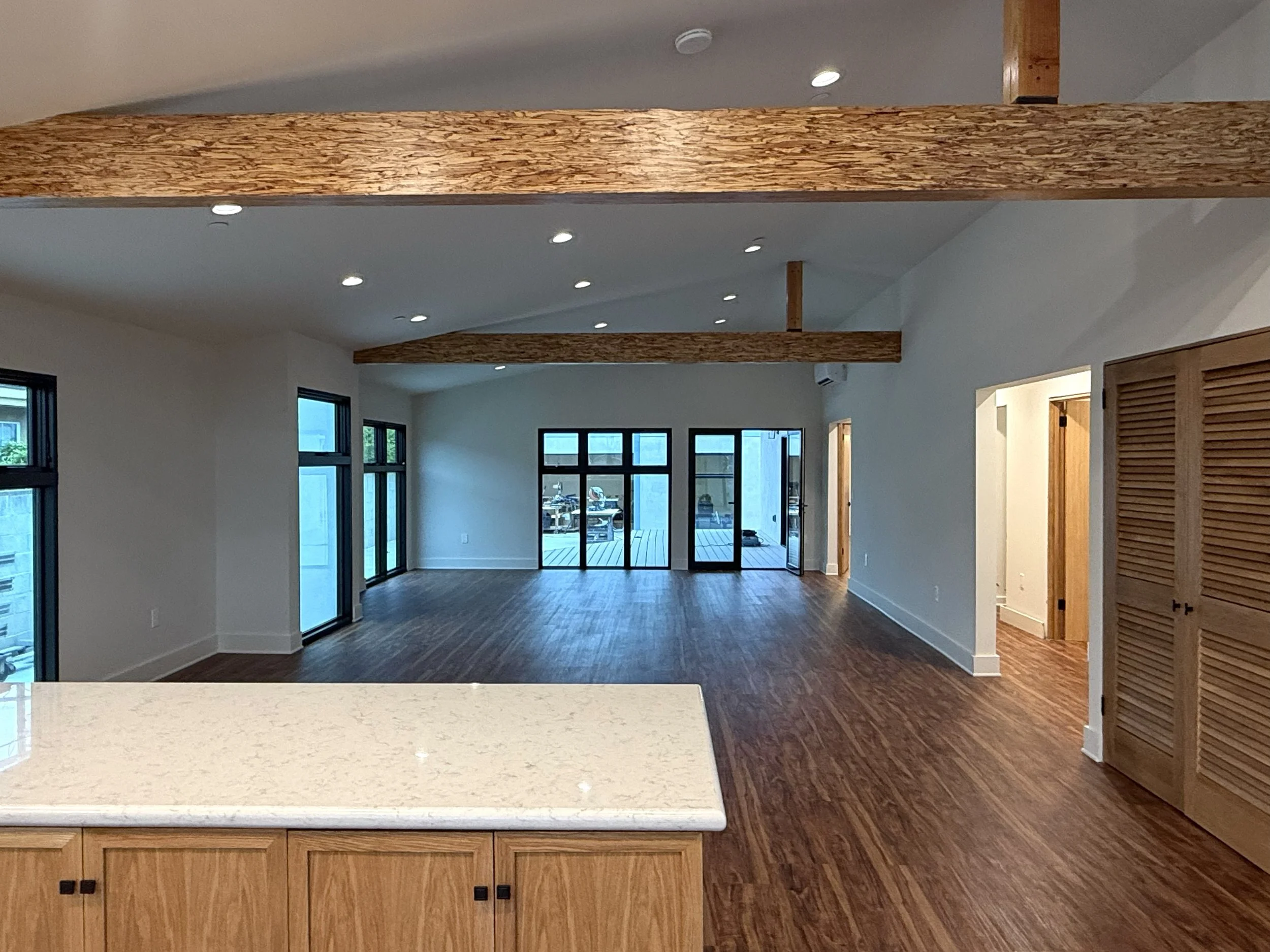 Empty living room with wooden floors, white walls, large windows, exposed wooden beams, and a sliding glass door leading outside.