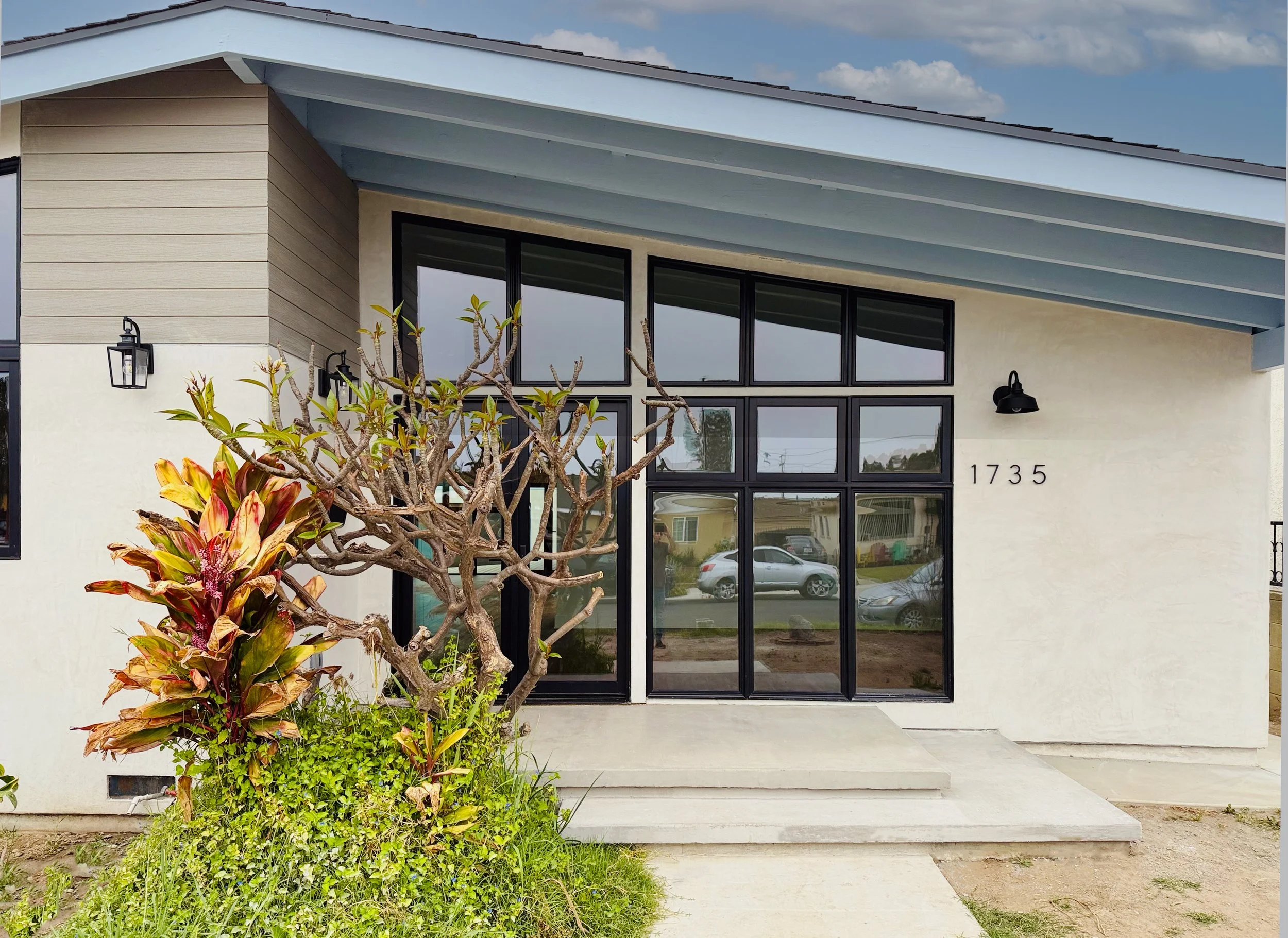 Front of a modern house with large black-framed windows, a concrete staircase leading to the entrance, a leafless tree, colorful plants, and the house number 1735 on the wall.