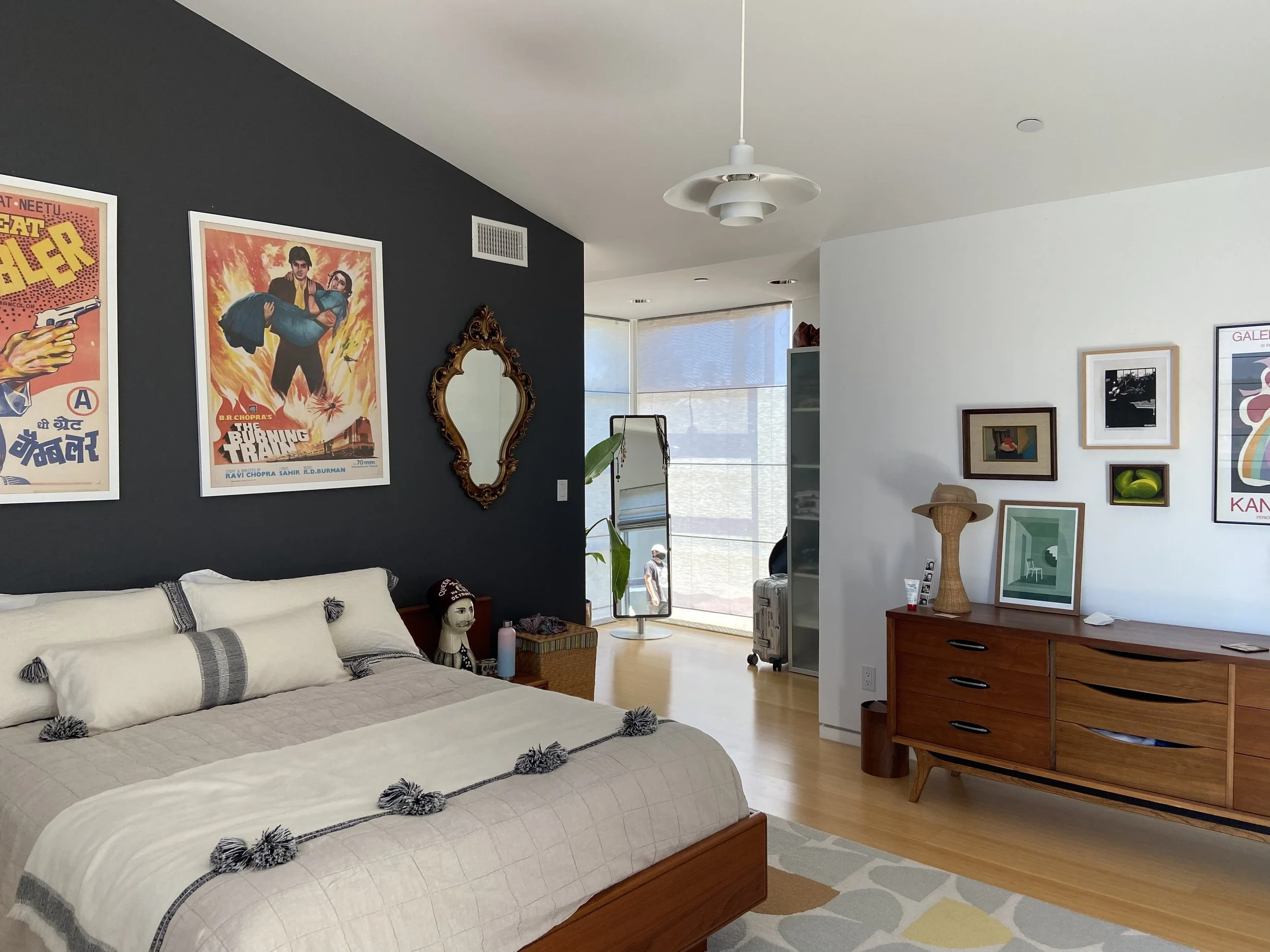 A bedroom with a white bed, dark accent wall with vintage posters, ornate mirror, and door leading outside. A wooden sideboard with artwork and a hat, and an open shelving unit near the window. Natural light streams in through partially open blinds.