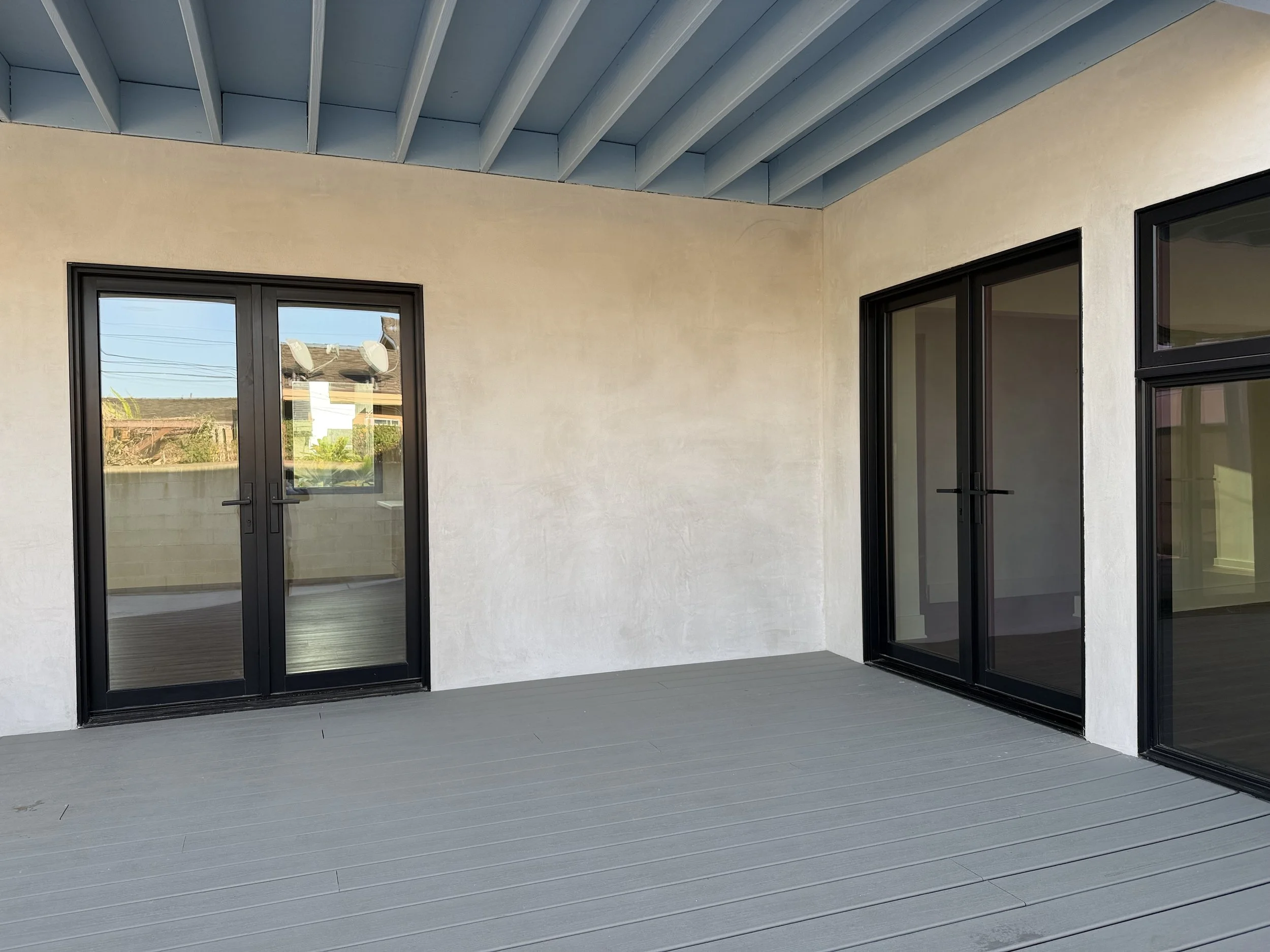A balcony with folding glass doors leading to an indoor space, with a textured beige wall and a blue ceiling with exposed beams.