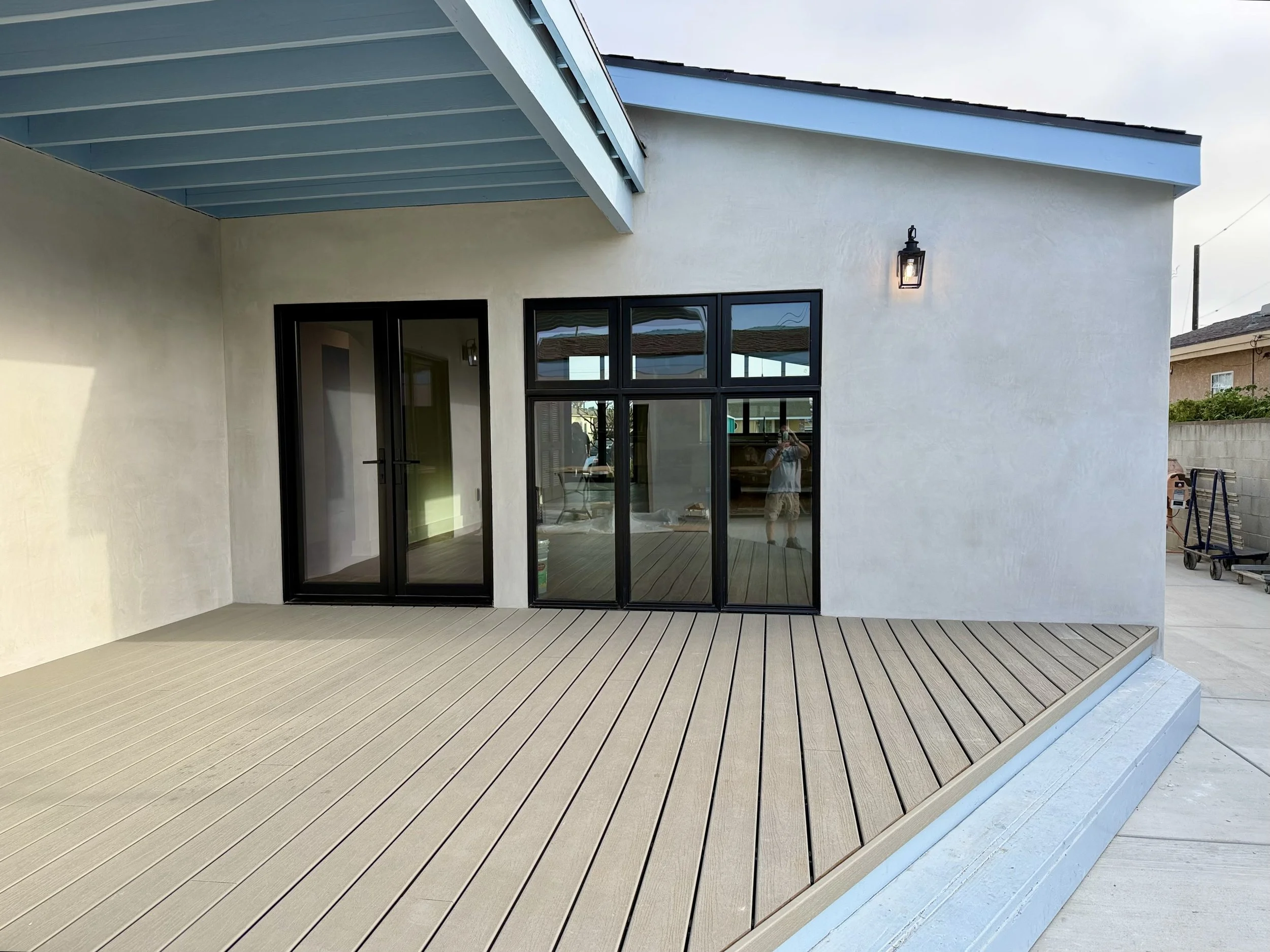 Newly built house with a beige wooden deck, black framed glass door, and window, exterior wall-mounted light