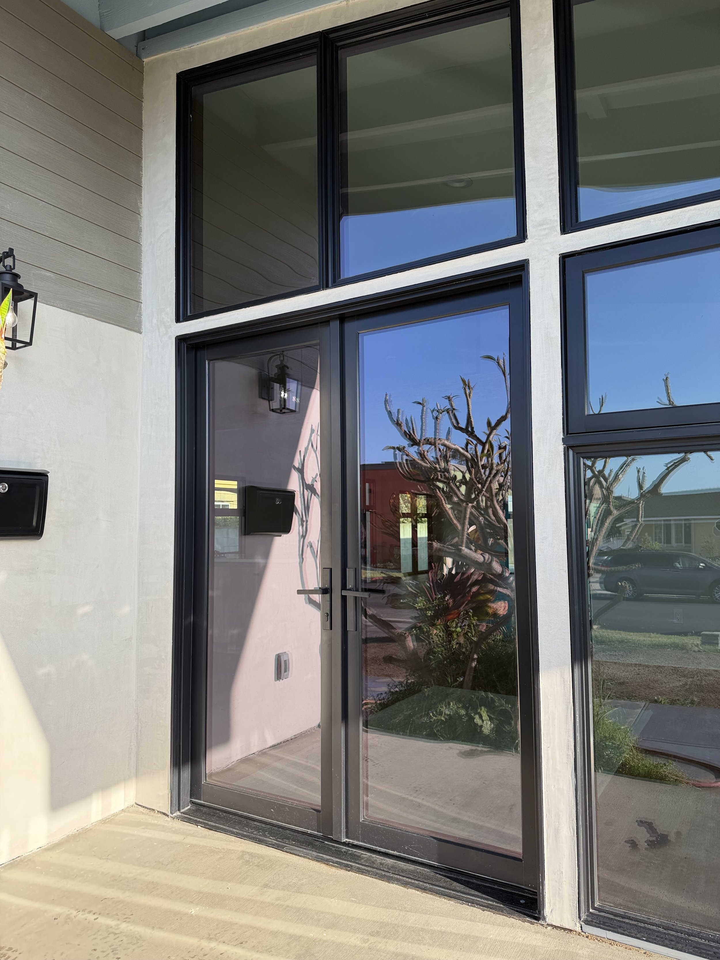 Front door of a modern house with glass panels, a black door handle, and reflection of a tree, blue sky, and neighboring houses.