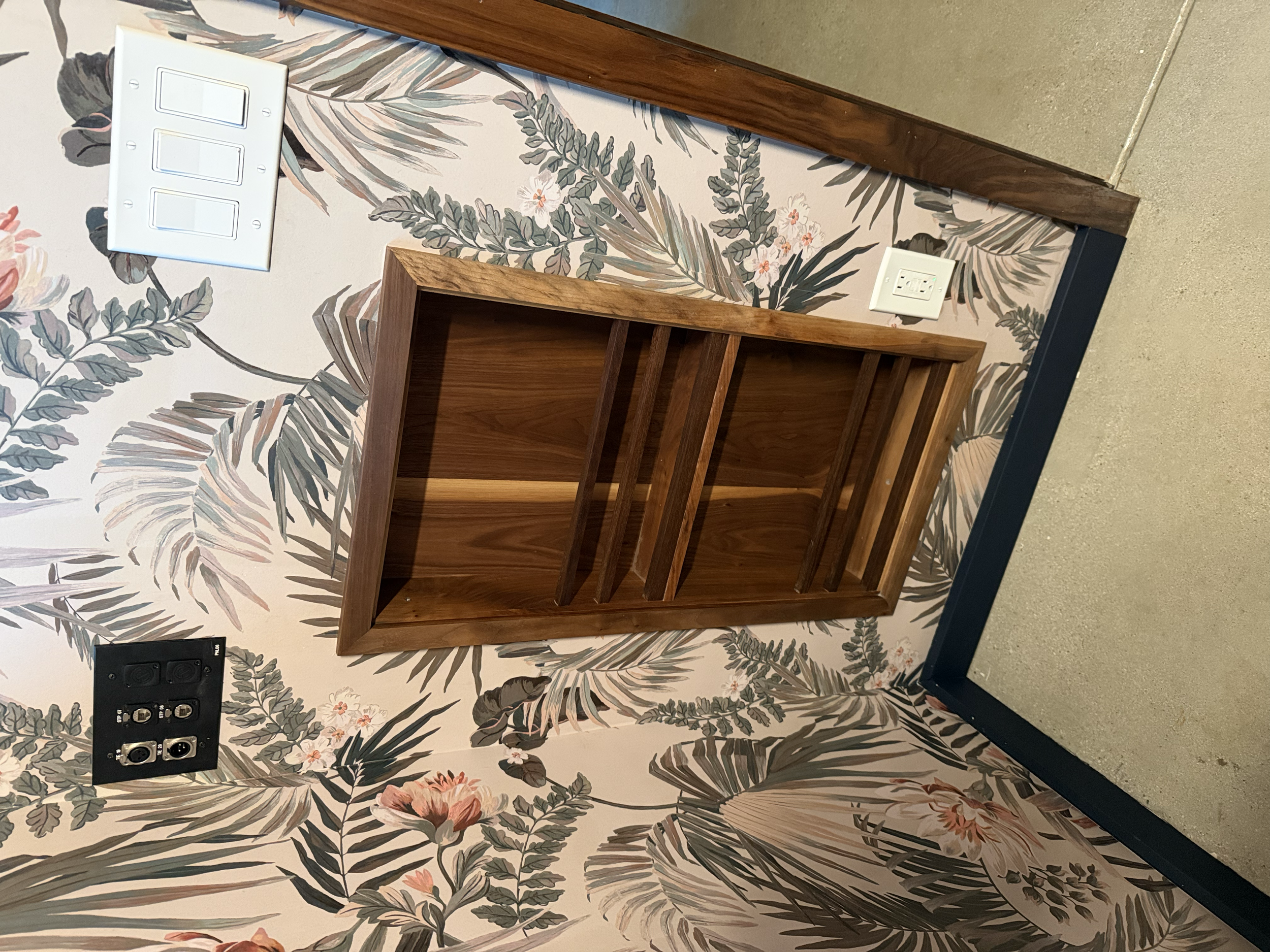 Empty wooden bookshelf mounted on a wall with a floral wallpaper background, electrical outlets, and switches nearby.