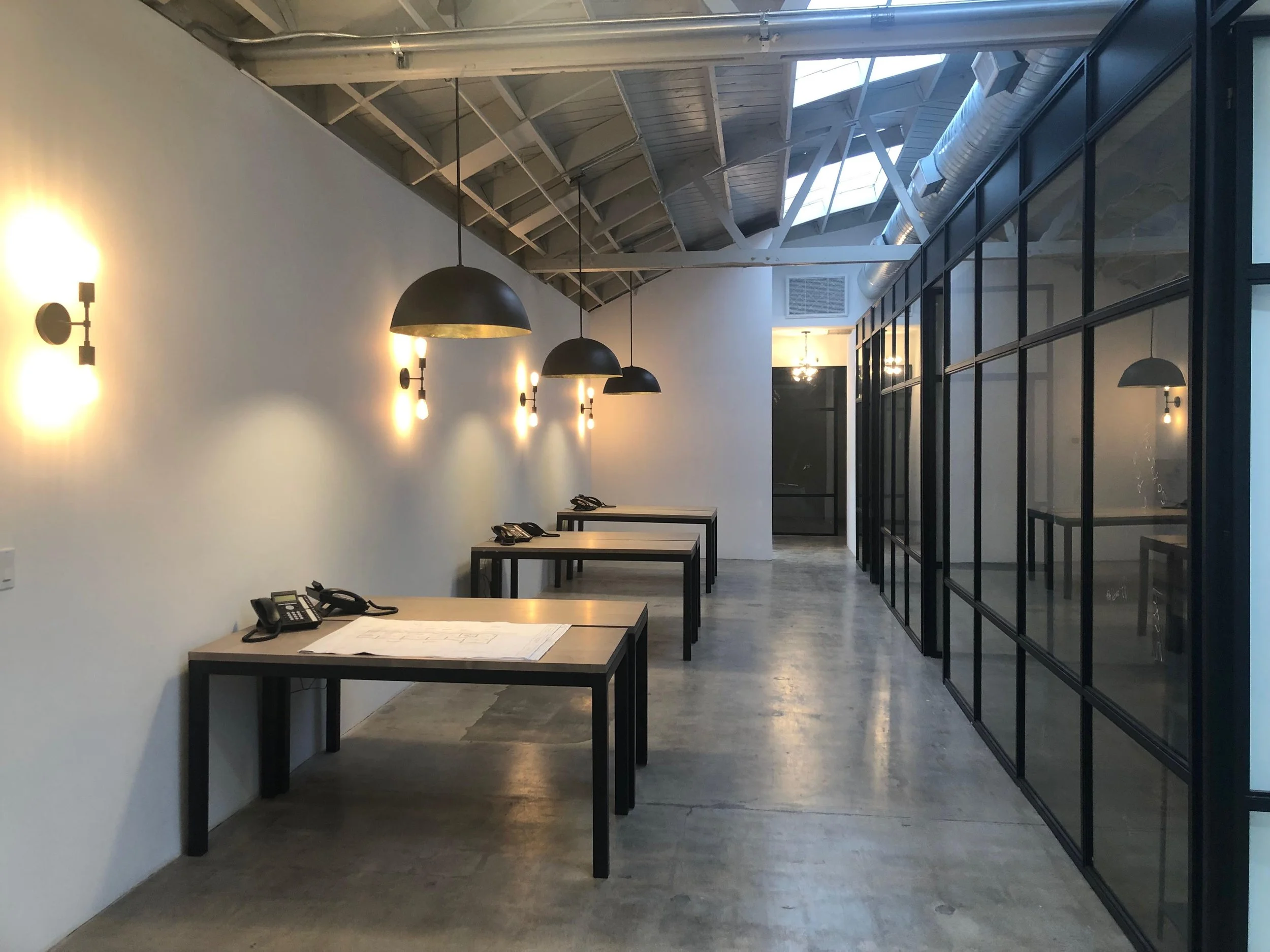 An empty modern office hallway with sunlight coming through a skylight. There are three desks with phones and papers, black pendant lights hanging overhead, and black-framed glass walls on the right side.