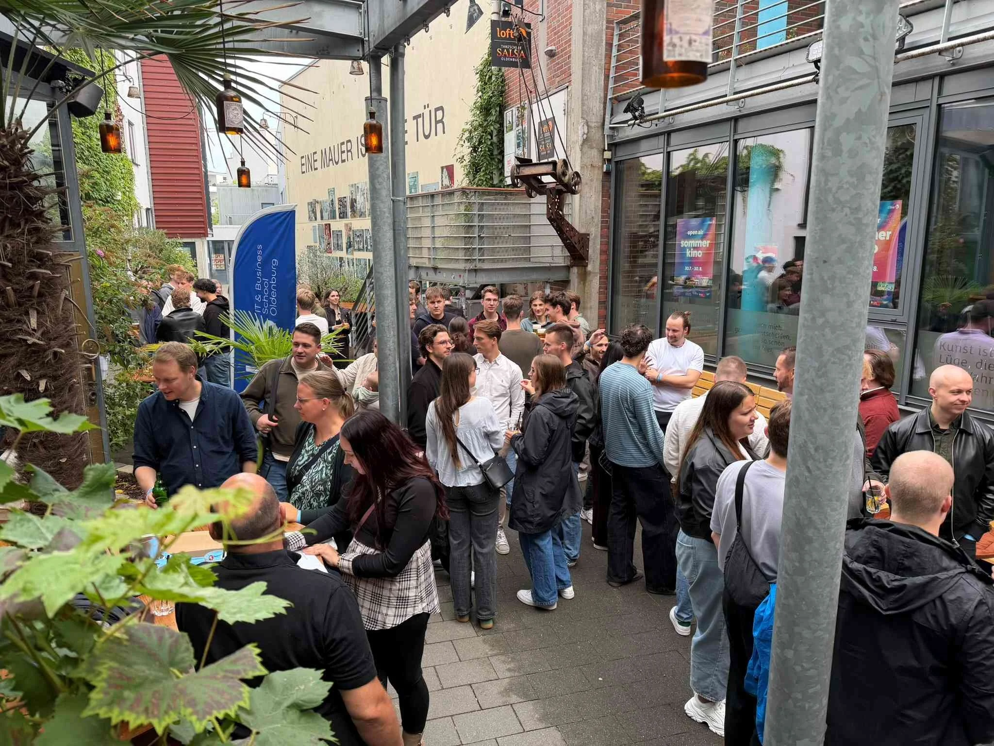 Gäste stehen in geselliger Runde unter einem überdachten Innenhof mit Glasdach und stimmungsvoller Beleuchtung beim Sommerfest der IBS Oldenburg.