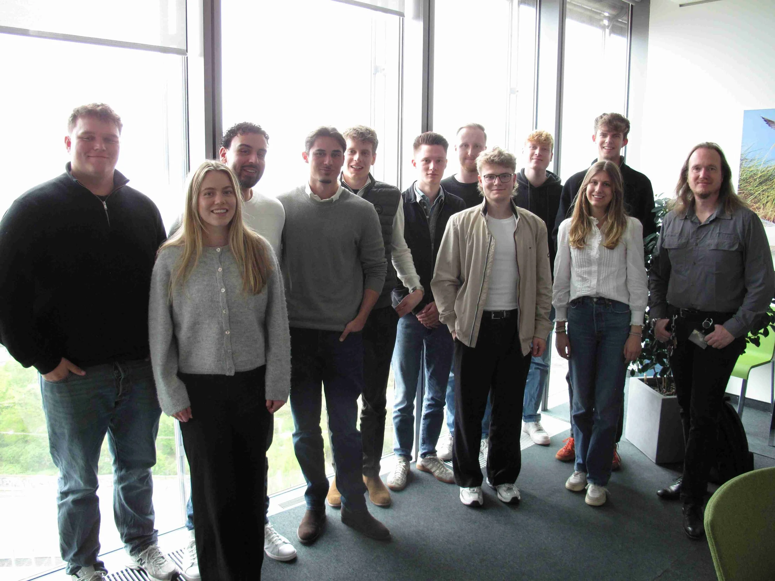 Eine Gruppe von Studierenden der IBS Oldenburg steht lächelnd für ein Gruppenfoto vor einer großen Fensterfront in einem modernen Bürogebäude während einer Exkursion.