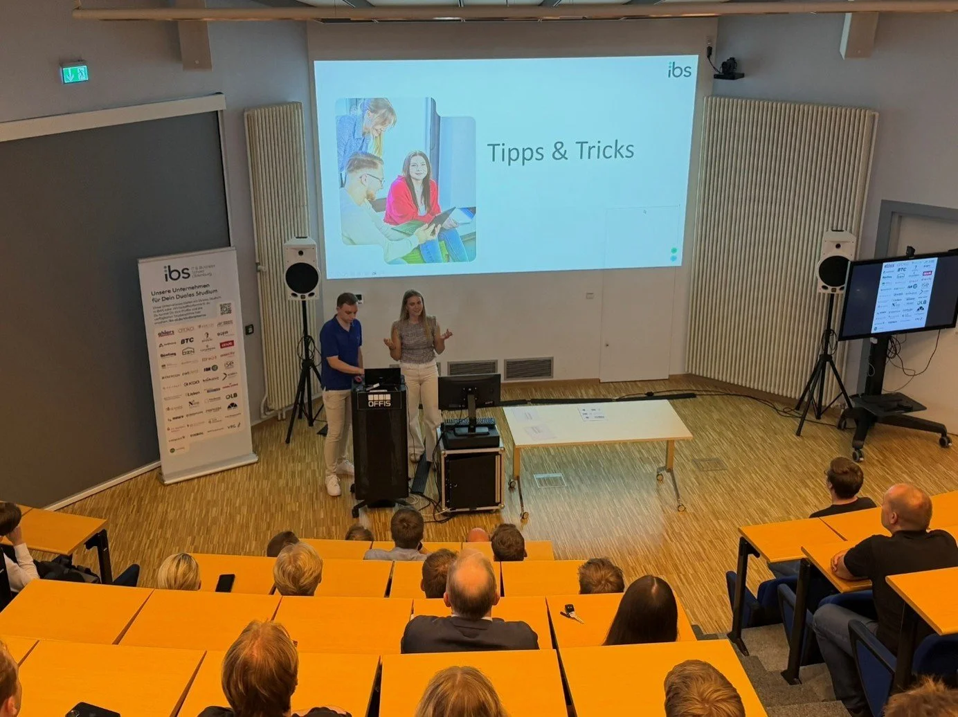 Zwei Personen präsentieren auf der Bühne eines Hörsaals das Thema „Tipps & Tricks“ auf einer Großleinwand vor Publikum an der IBS Oldenburg.