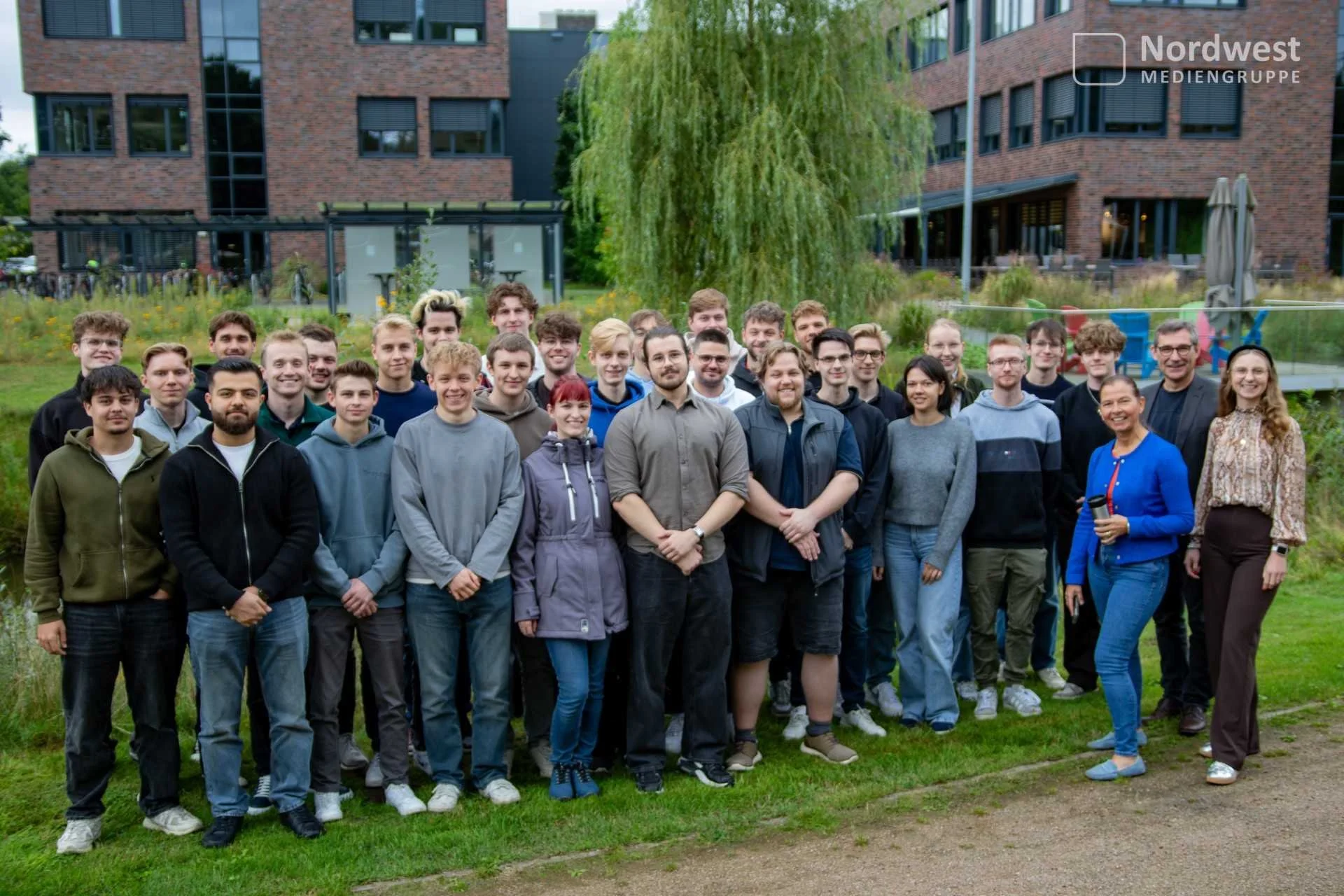 Eine große Gruppe junger Menschen und Vertretender der Nordwest Mediengruppe steht für ein Gruppenfoto auf einer Grünfläche vor modernen Klinkergebäuden.
