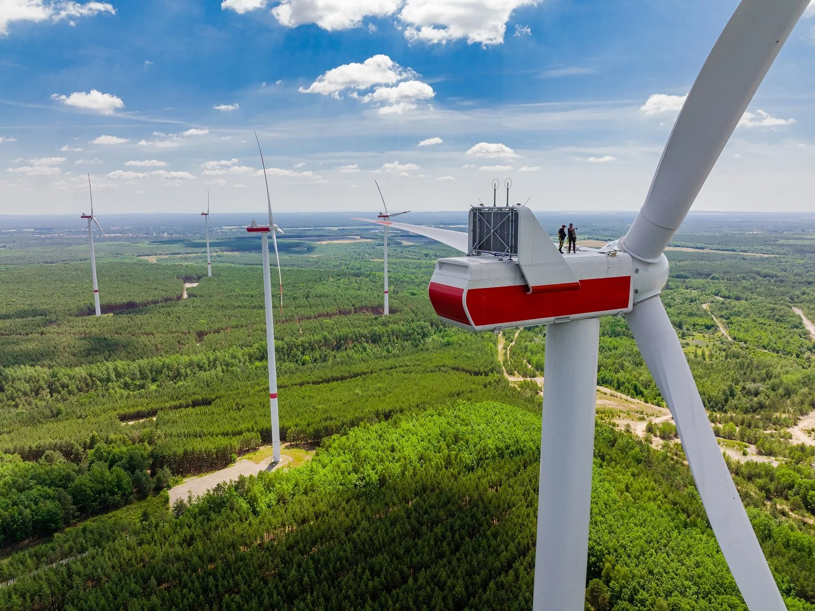Luftaufnahme eines Windparks in einem Waldgebiet. Zwei Techniker stehen auf der Gondel einer rot-weißen Windkraftanlage der enercity AG unter strahlend blauem Himmel.