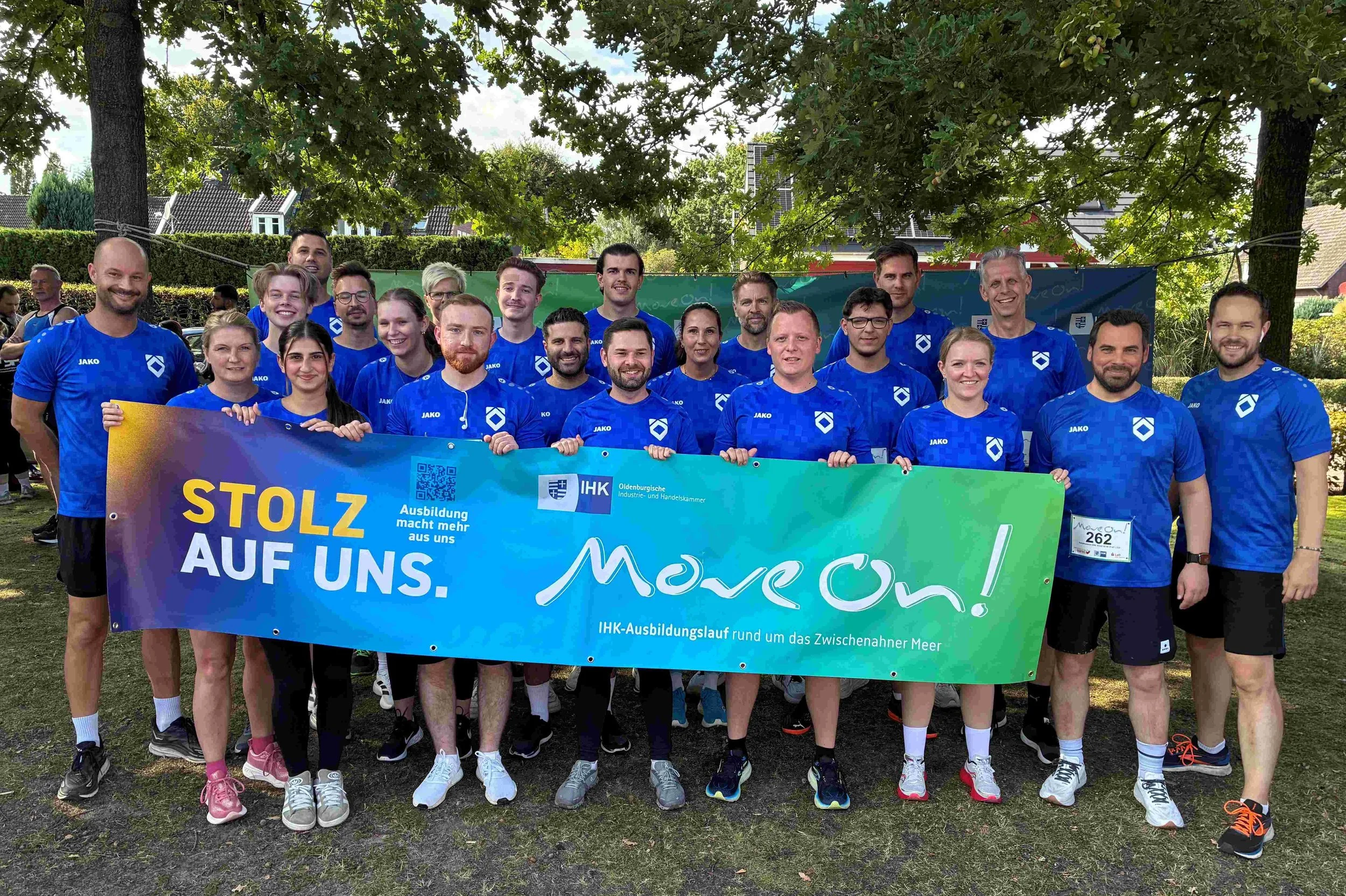 Ein Team in blauen Trikots hält ein Banner zum IHK-Ausbildungslauf „Move On!“ am Zwischenahner Meer hoch. Text: „STOLZ AUF UNS. Ausbildung macht mehr aus uns.“.