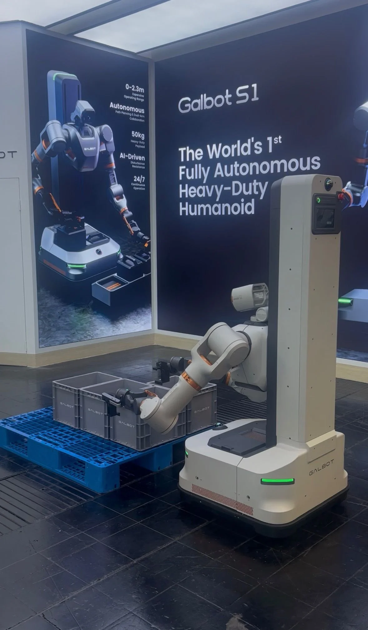 Ein Galbot S1 Roboter auf einer Messe bei einer Demonstration. Der autonome humanoide Roboterarm greift nach grauen Boxen auf einer blauen Palette vor einer Infowand.