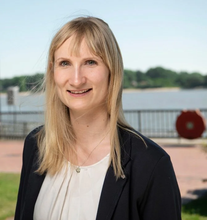 Porträt von Thekla, einer blonden Frau im dunklen Blazer, die lächelnd im Freien steht. Im Hintergrund sind eine Uferpromenade und ein Gewässer bei sonnigem Wetter erkennbar.