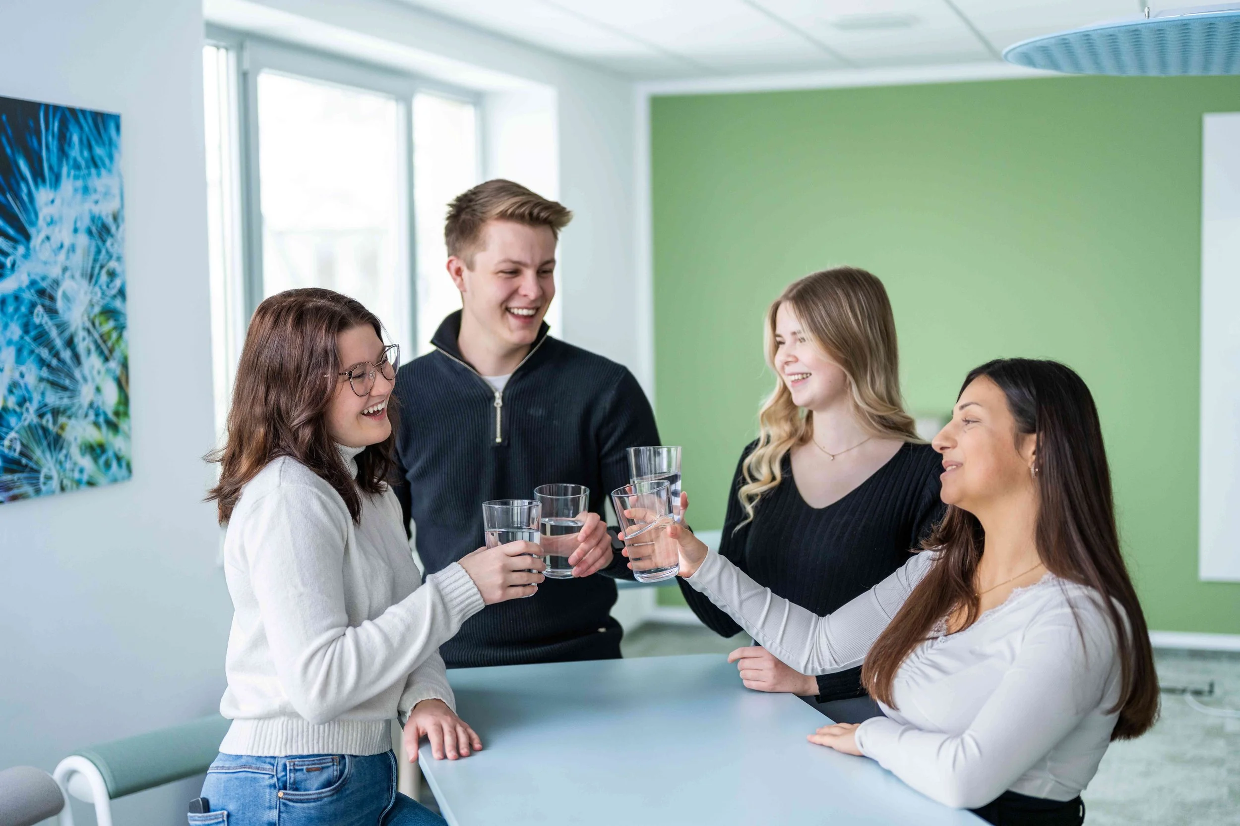 Vier junge Leute stehen lachend an einem hohen Tisch in einem modernen Raum und stoßen mit Wassergläsern an. Die Atmosphäre ist gelöst und freundlich