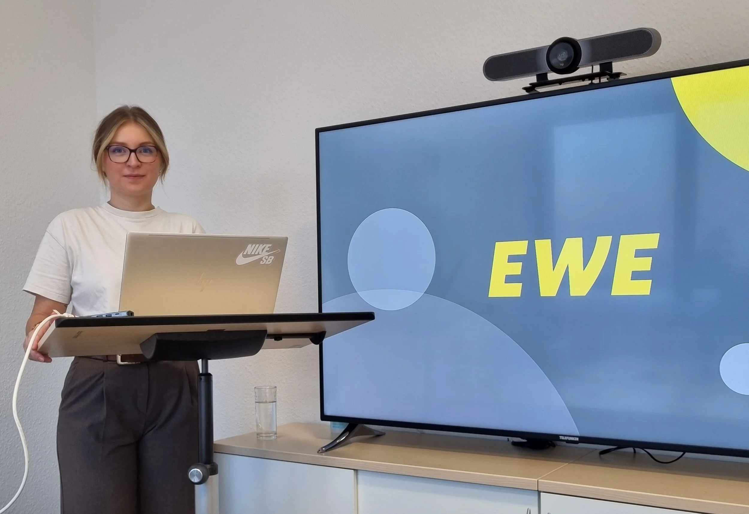 Eine Studentin steht mit einem Laptop an einem Stehpult neben einem großen Bildschirm mit dem „EWE“-Logo. Sie trägt eine Brille und bereitet sich offensichtlich auf eine Präsentation vor.