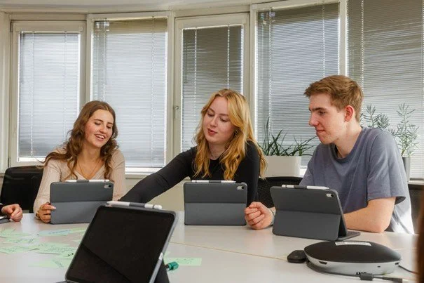 Drei Studierende sitzen an einem Tisch und arbeiten gemeinsam mit Tablets und Stylus-Stiften. Vor ihnen liegen Notizzettel; die Atmosphäre wirkt konzentriert und kollaborativ.