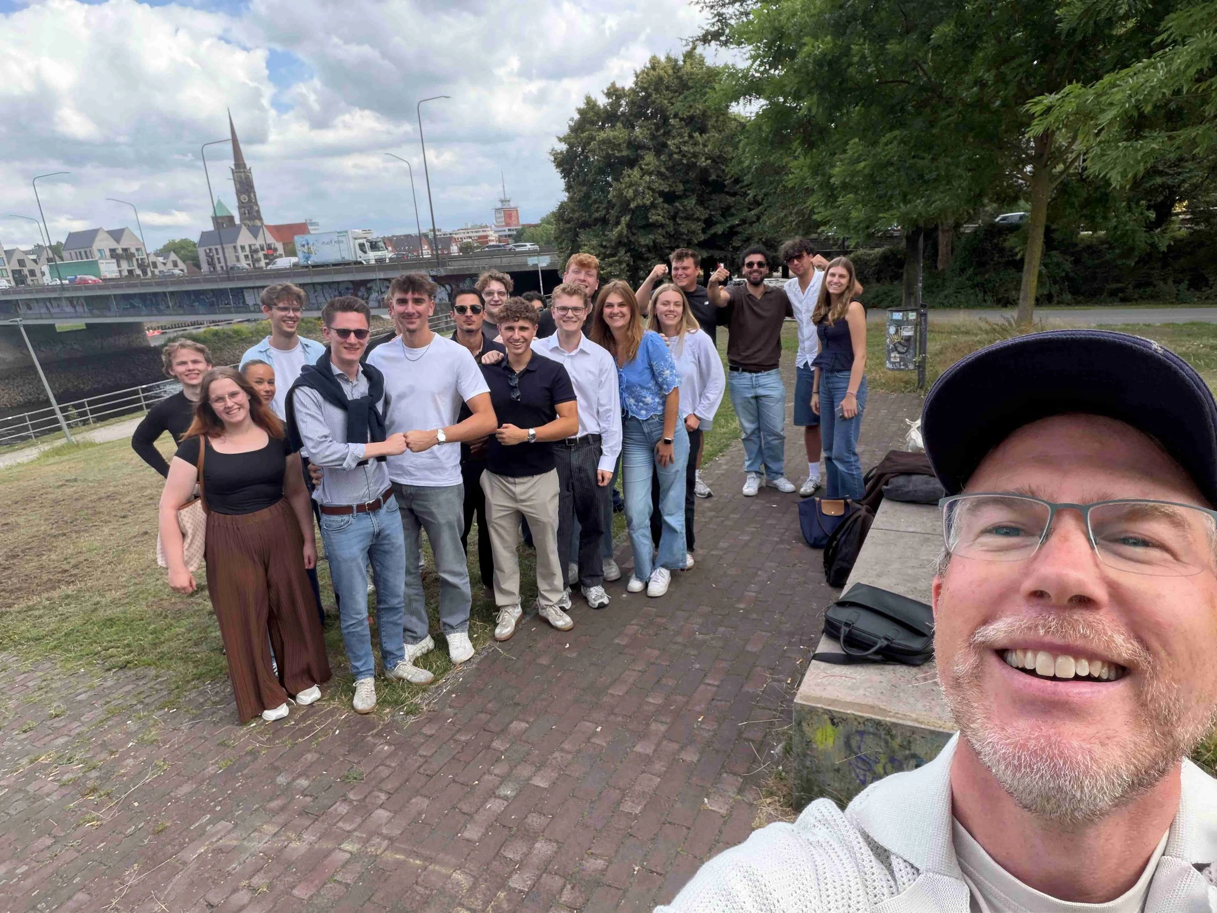 Ein lächelnder Mann macht ein Selfie mit einer großen Gruppe junger Leute im Freien vor einer Flusskulisse mit Brücke und Kirchturm im Hintergrund.