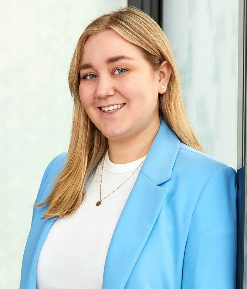 Porträtaufnahme einer lächelnden jungen Frau mit blonden Haaren. Sie trägt einen hellblauen Blazer über einem weißen T-Shirt und lehnt an einer Glaswand.