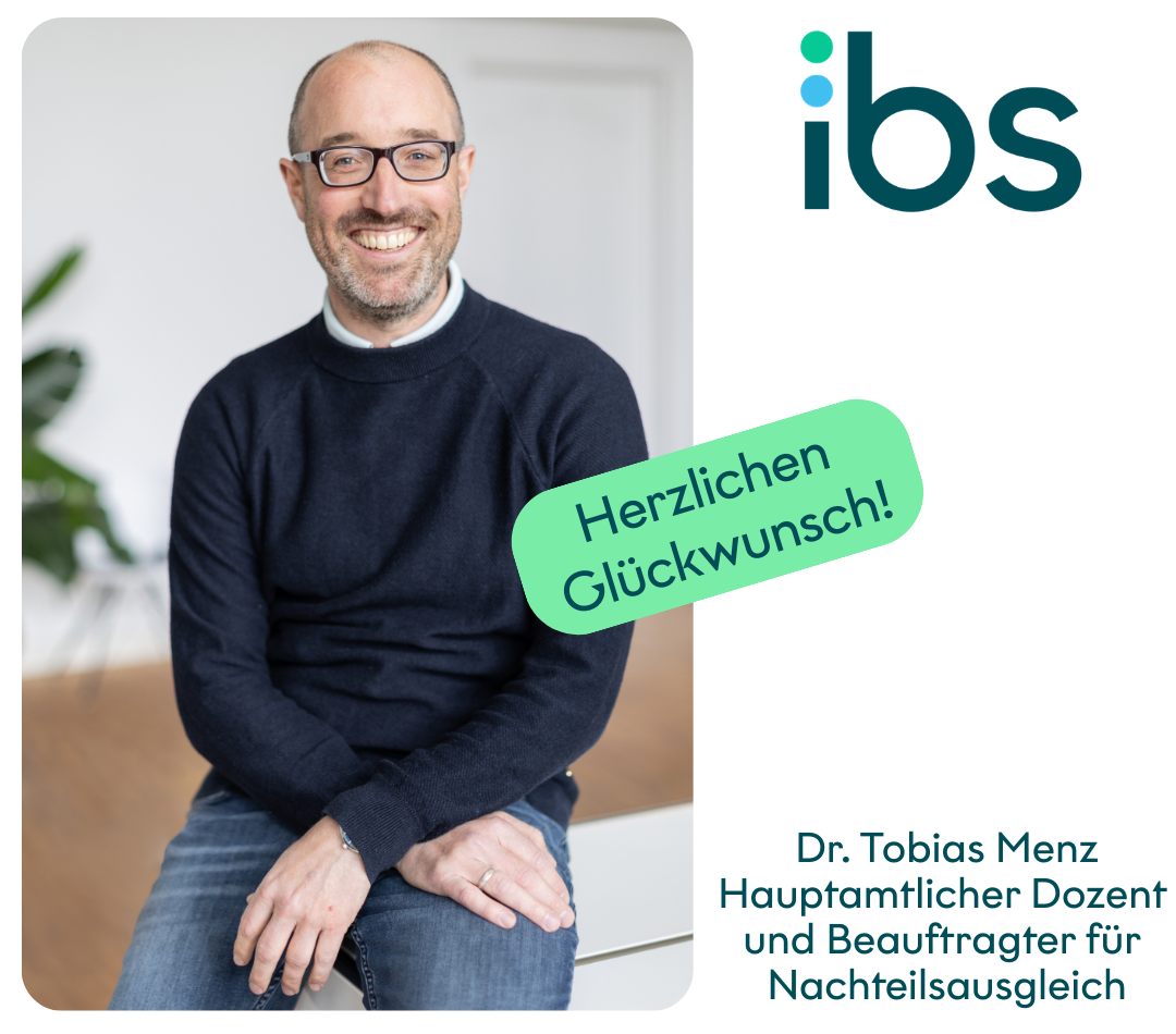 Das Bild zeigt Dr. Tobias Menz, Dozent an der IBS Oldenburg. Er sitzt lächelnd auf einer Tischkante, daneben ein Banner mit „Herzlichen Glückwunsch!“.
