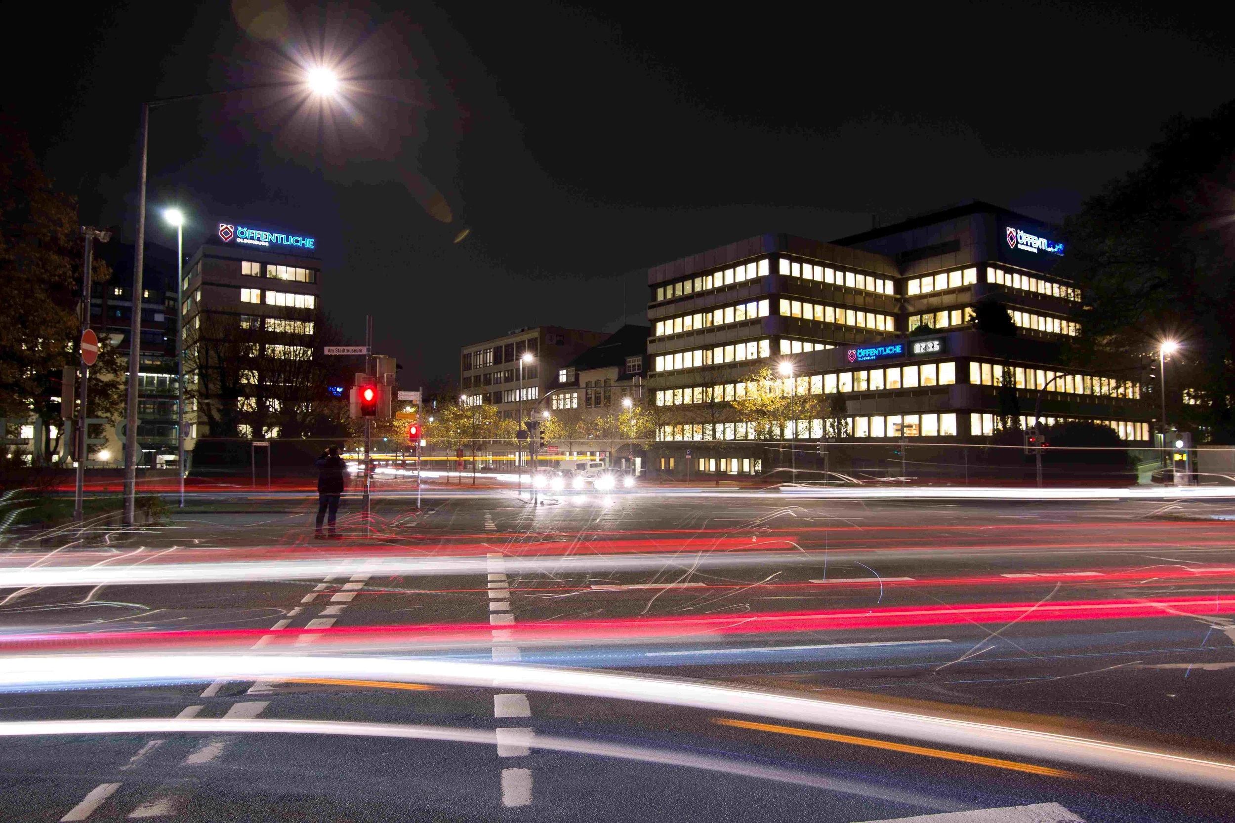 Beleuchtete Bürogebäude der Öffentlichen Oldenburg bei Nacht an einer Kreuzung. Langzeitbelichtung erzeugt dynamische Lichtspuren von Autos im Vordergrund unter einem dunklen Nachthimmel.