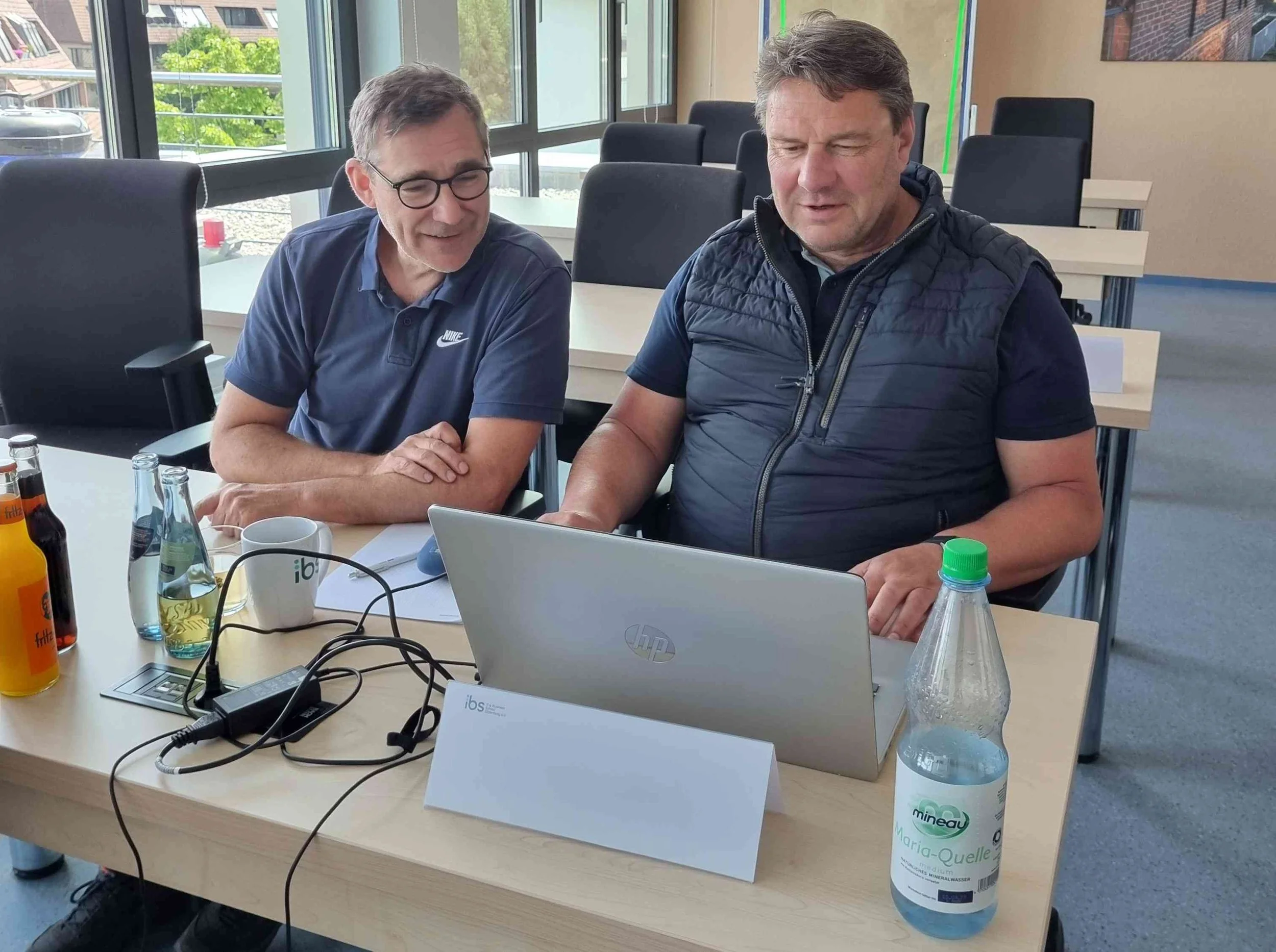 Zwei Lehrende der IBS Oldenburg sitzen nebeneinander an einem Tisch im Seminarraum und blicken konzentriert auf den Bildschirm eines geöffneten Laptops.