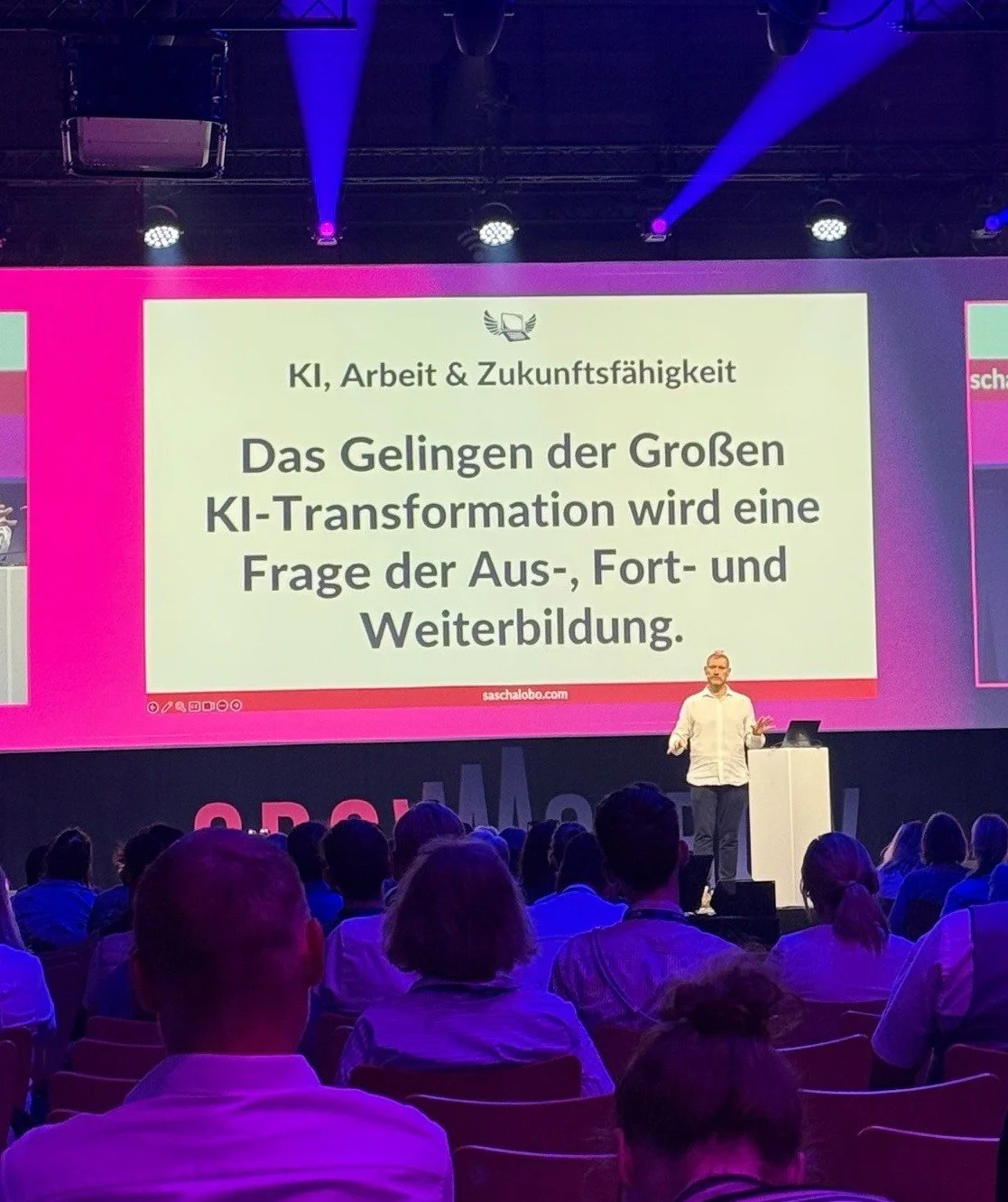 Sascha Lobo hält auf einer Bühne einen Vortrag zum Thema KI. Im Hintergrund zeigt eine Leinwand die Aufschrift: „Das Gelingen der Großen KI-Transformation wird eine Frage der Aus-, Fort- und Weiterbildung.“