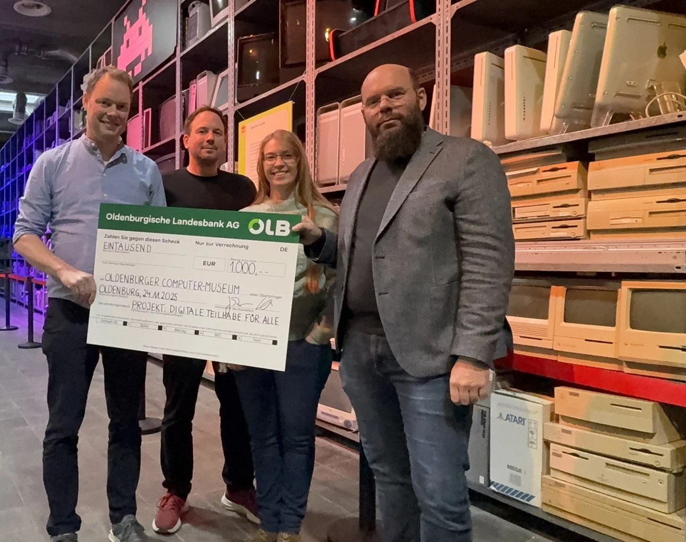 Vier Personen stehen in einem Computermuseum und halten einen Spendenscheck der OLB über 1.000 € für das Projekt „Digitale Teilhabe für alle“. Im Hintergrund sind alte Computer in Regalen.