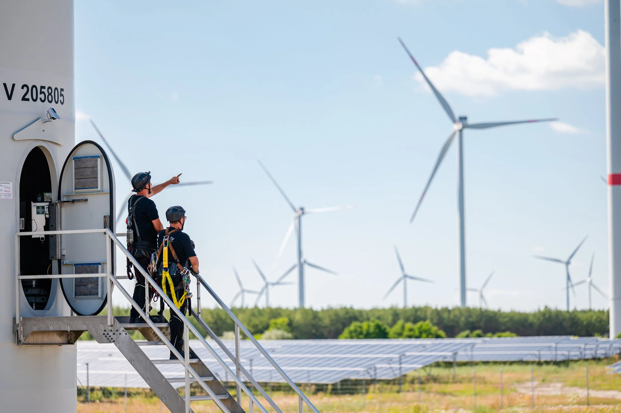 Zwei Techniker in Schutzausrüstung stehen auf der Außentreppe einer Windkraftanlage. Im Hintergrund erstreckt sich ein Windpark über einer großen Photovoltaikanlage bei sonnigem Wetter.