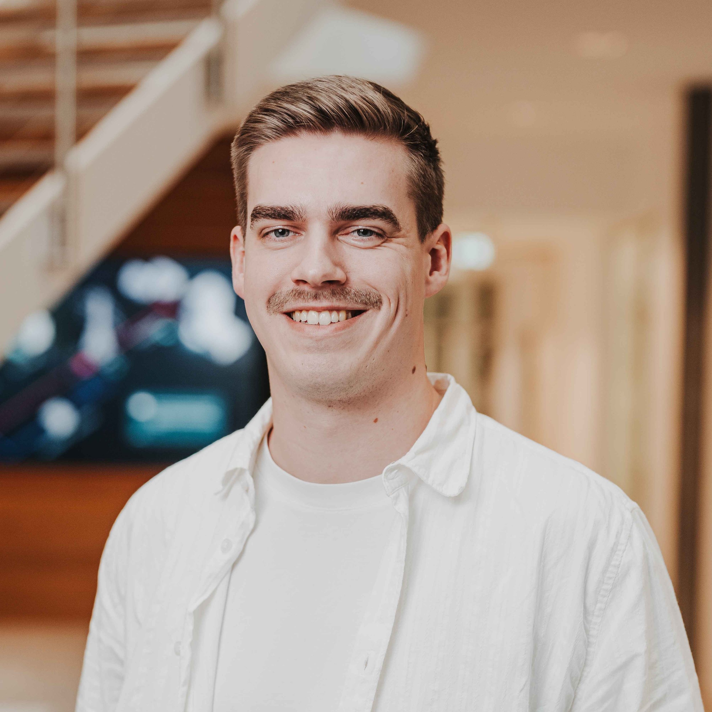 Ein lächelnder junger Mann mit kurzen braunen Haaren und Schnurrbart. Er trägt ein weißes Hemd über einem T-Shirt vor einem unscharfen, hellen Innenraum-Hintergrund mit Treppenansatz.