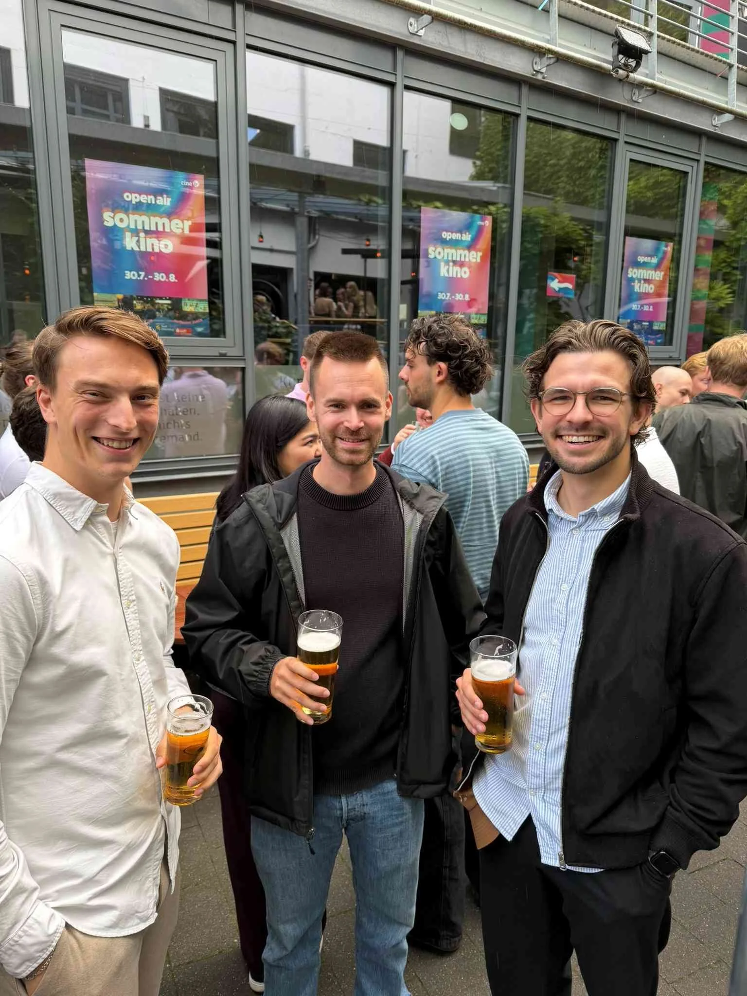 Drei lächelnde junge Männer stehen mit Getränken im Freien vor Fenstern mit Veranstaltungspostern beim Sommerfest der IBS Oldenburg.