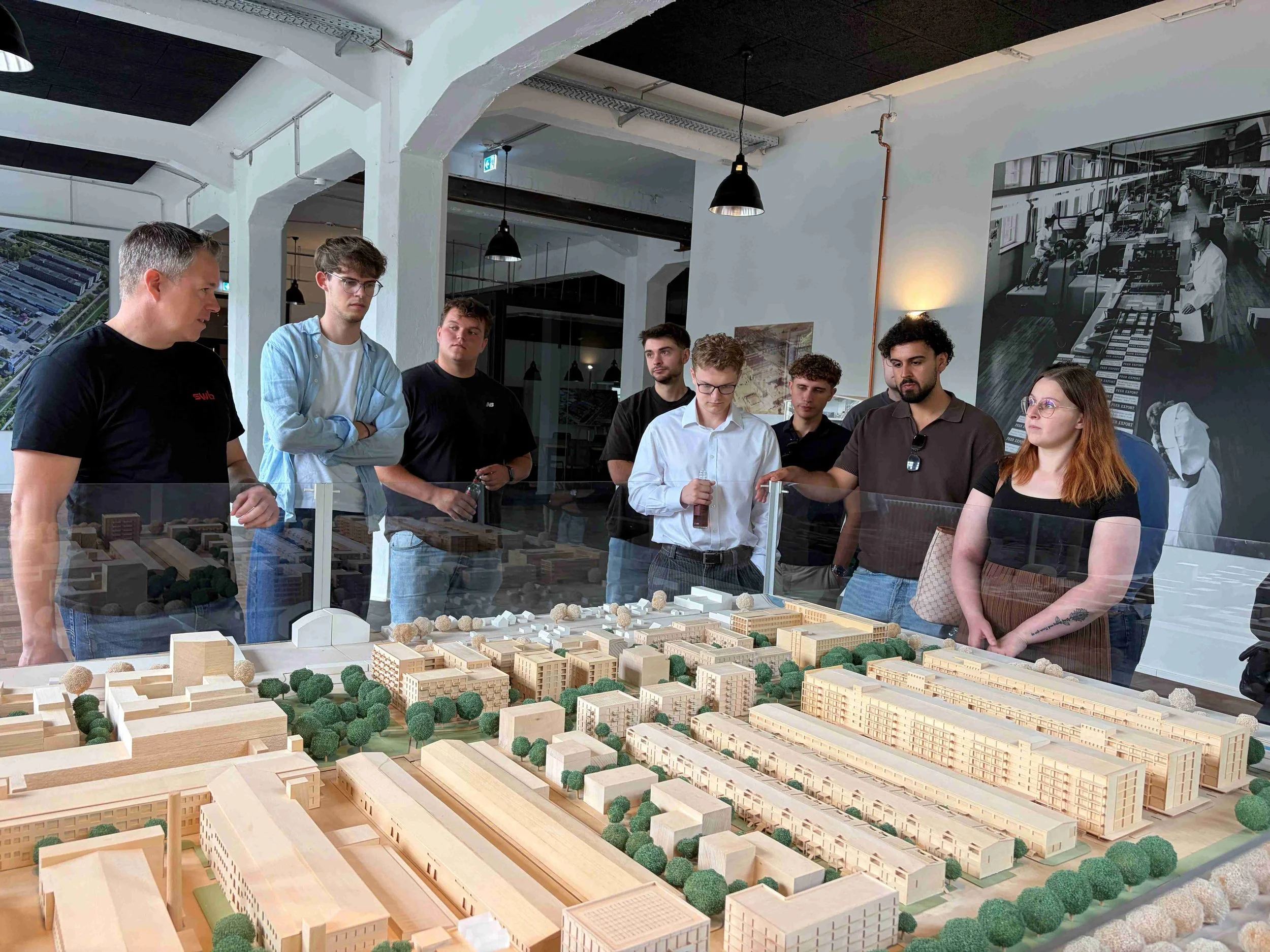 Eine Gruppe junger Leute steht auf einer Exkursion aufmerksam um ein großes, detailliertes Architekturmodell aus Holz herum und lässt sich die Stadtplanung erklären.