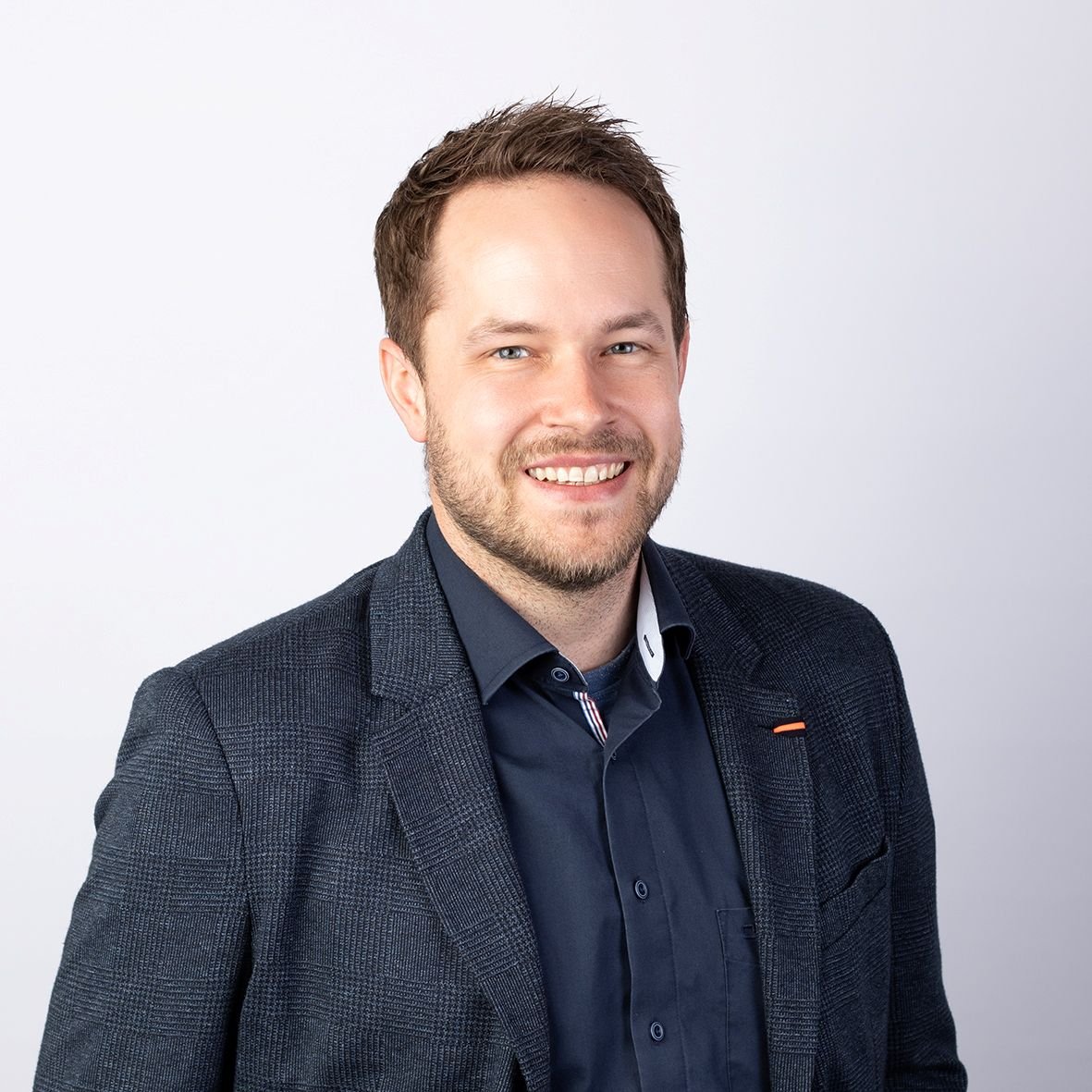 Ein lächelnder Mann mit kurzen braunen Haaren und Bart blickt direkt in die Kamera. Er trägt ein schickes, dunkelblaues Sakko über einem blauen Hemd vor einem hellen, schlichten Hintergrund.