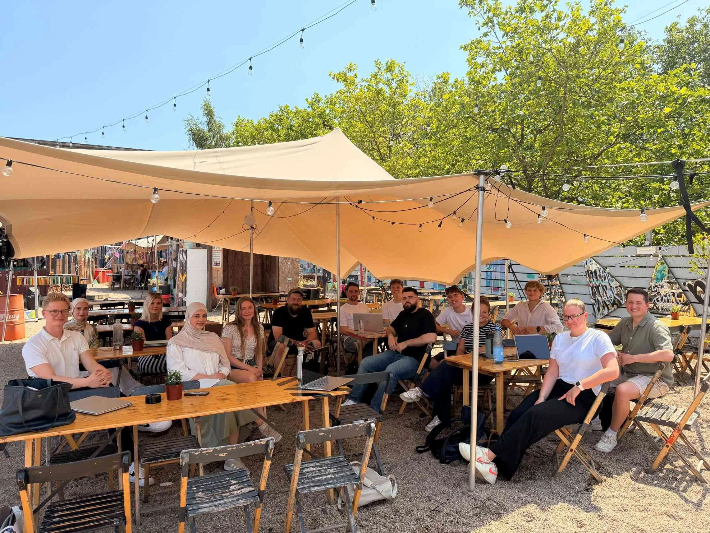 Eine Gruppe von Studierenden der IBS Oldenburg sitzt während einer Exkursion gut gelaunt an Holztischen unter einem großen Sonnensegel in einem gemütlichen Outdoor-Bereich.