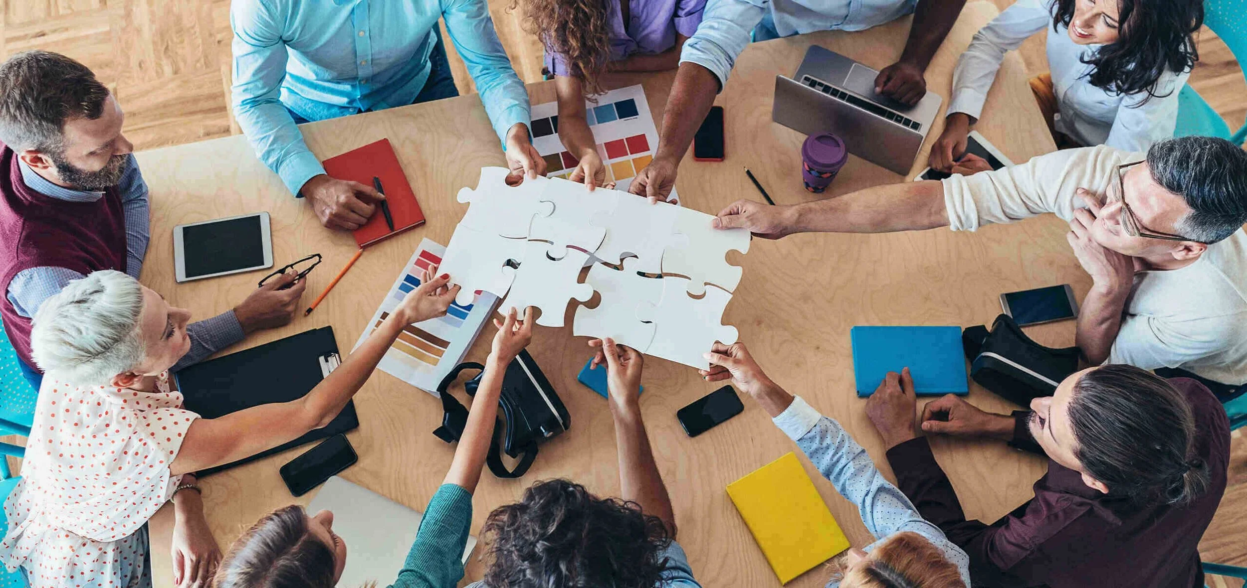 Draufsicht auf ein Team an einem Holztisch, das gemeinsam große weiße Puzzleteile zusammensetzt. Ein Symbolbild für Kooperation, Teamwork und Problemlösung im Studium.